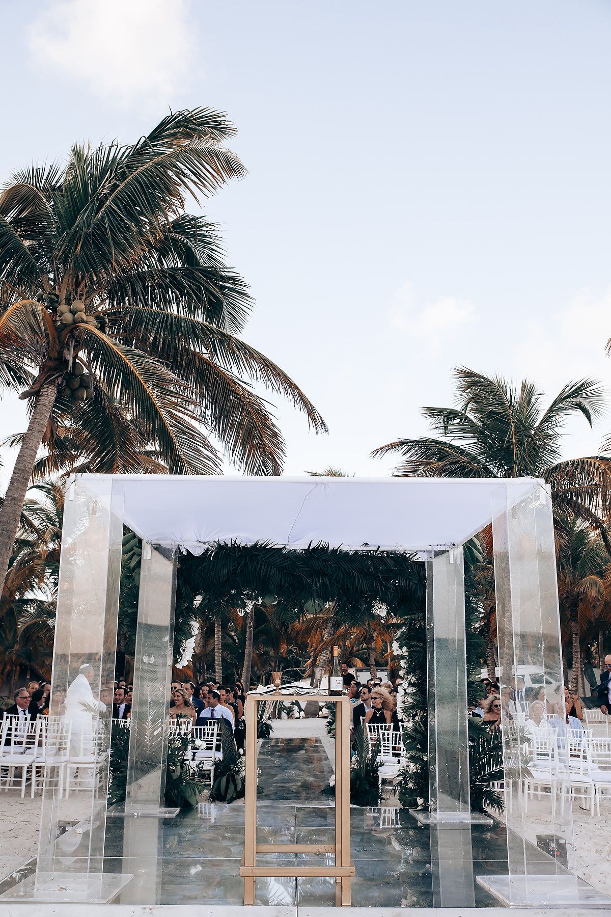 Tropical beach wedding ceremony setup with white canopy and chairs under palm trees.