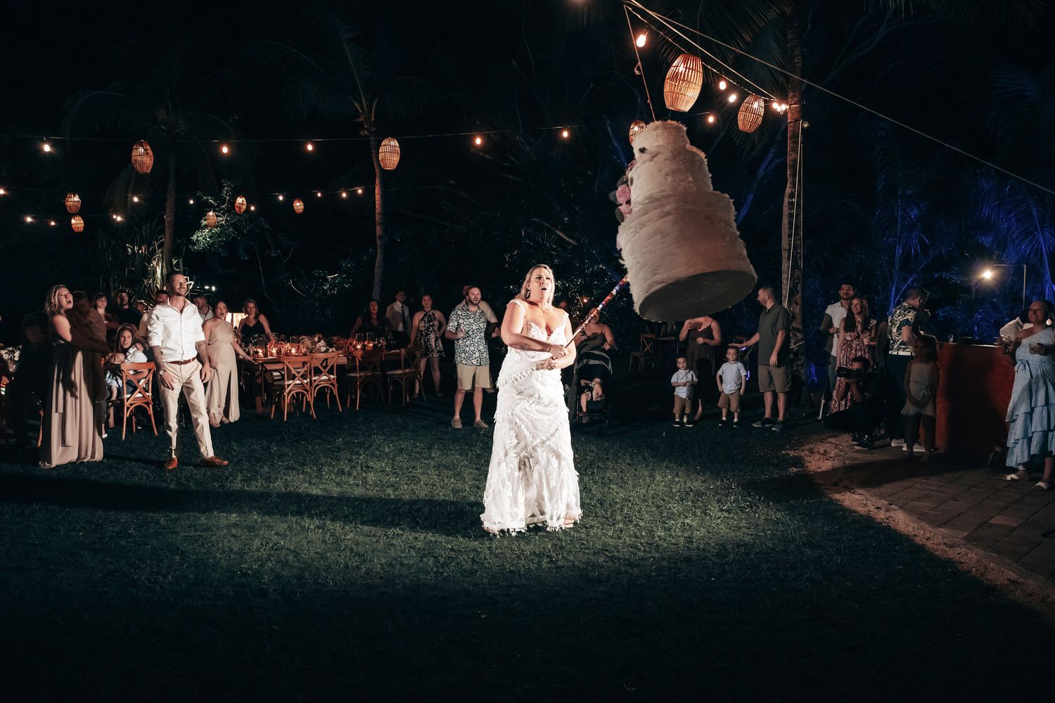 Bride in lace wedding dress stands alone on grass at elegant outdoor nighttime wedding reception.