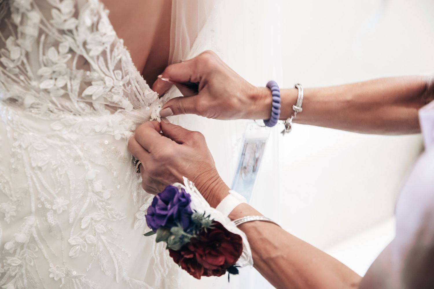 Close-up of hands fastening buttons on intricate lace wedding dress with floral corsage.