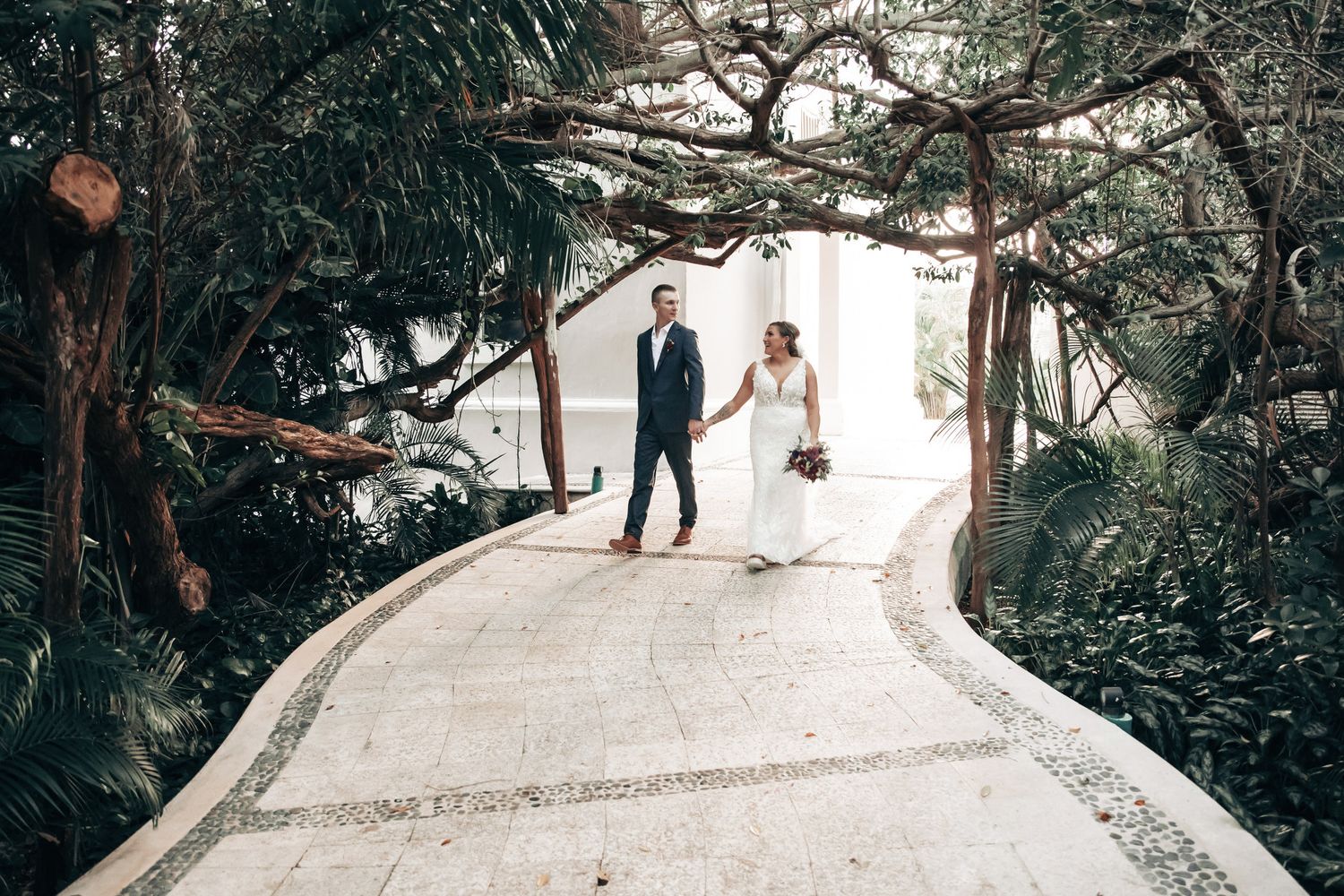 Bride and groom walking hand in hand on elegant garden pathway lined with lush trees.