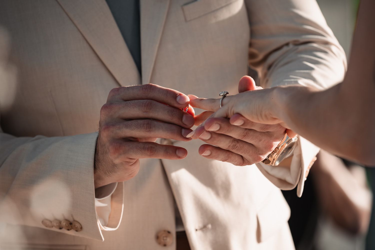 Close-up of groom placing wedding ring on bride's finger during outdoor ceremony in beige suit.