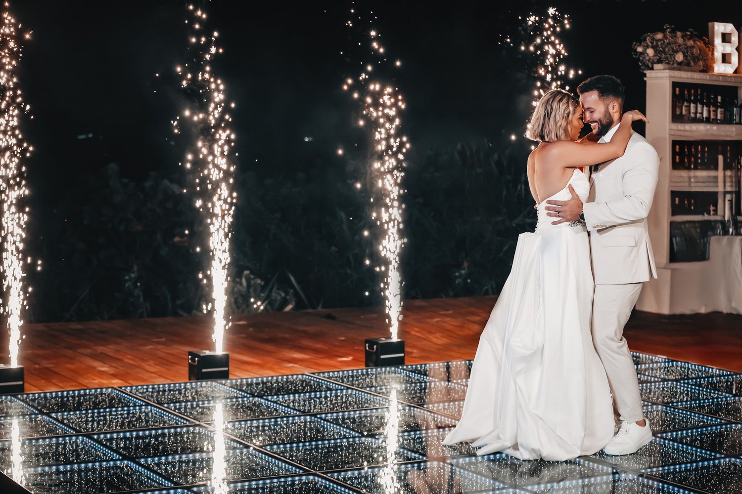 Bride and groom first dance on mirrored floor with sparkling cold firework fountains at night.