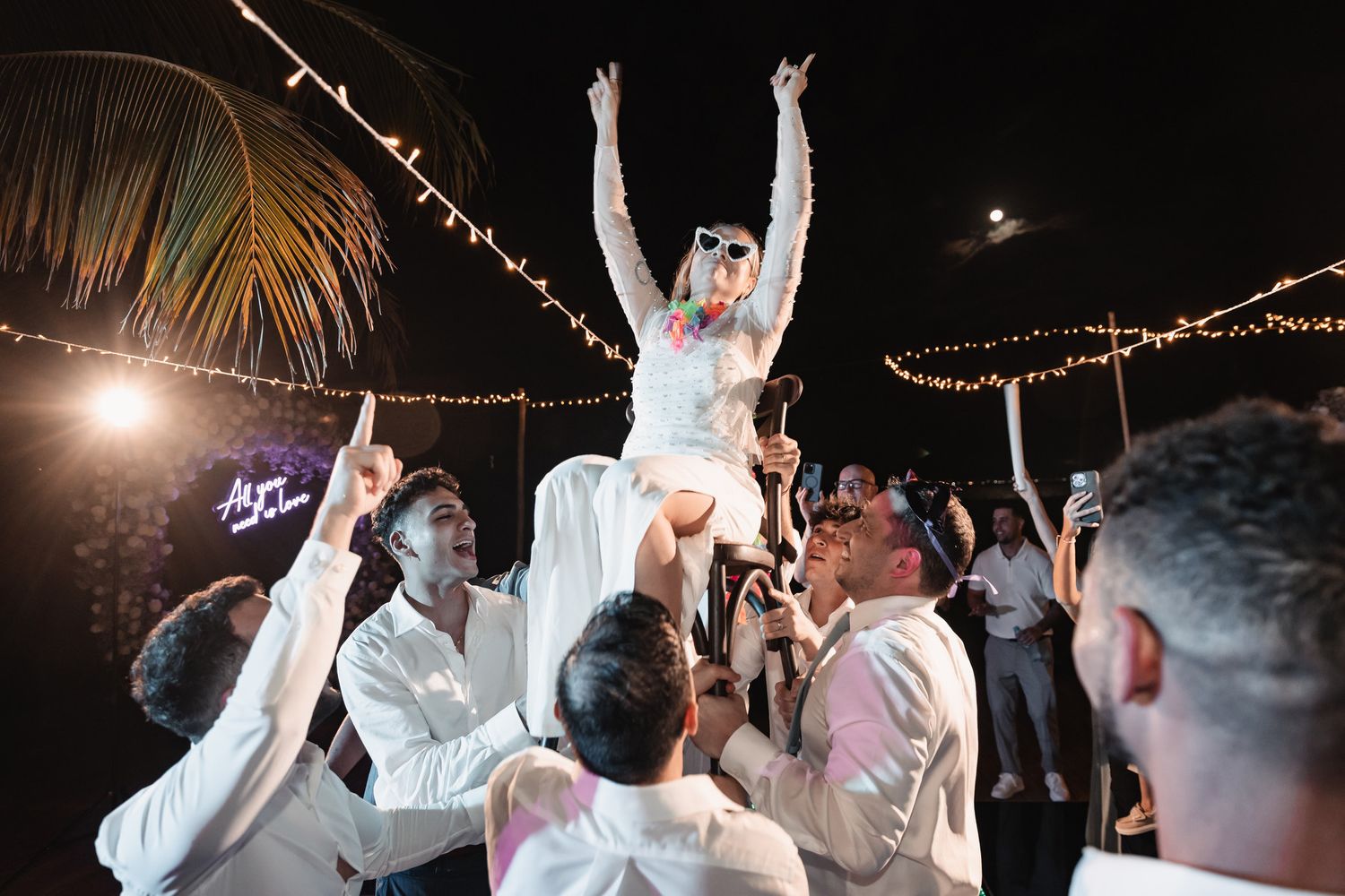 Bride raised up by groomsmen during lively outdoor wedding reception celebration at night.