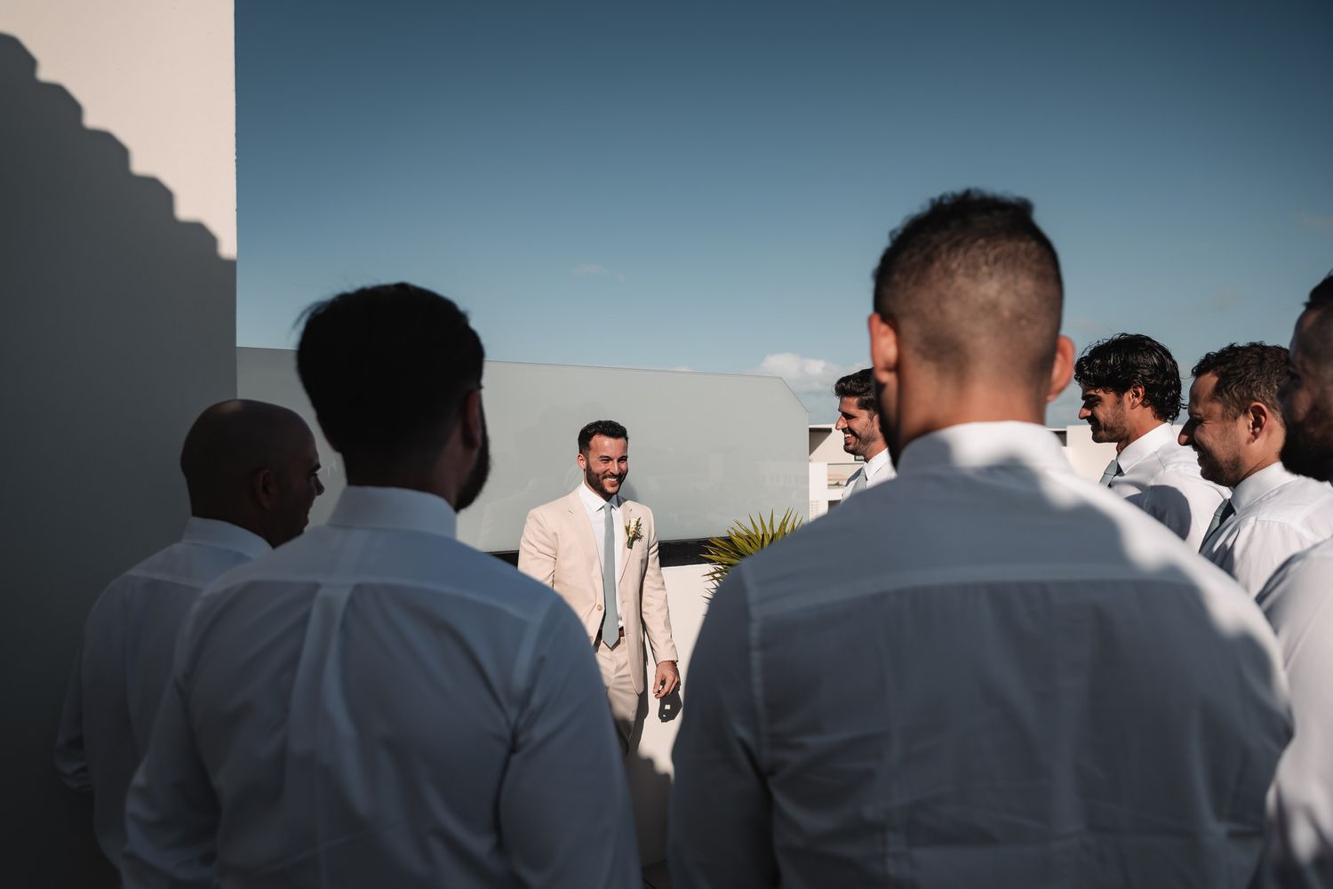 Groom in beige suit greeting groomsmen in white shirts on outdoor rooftop venue.