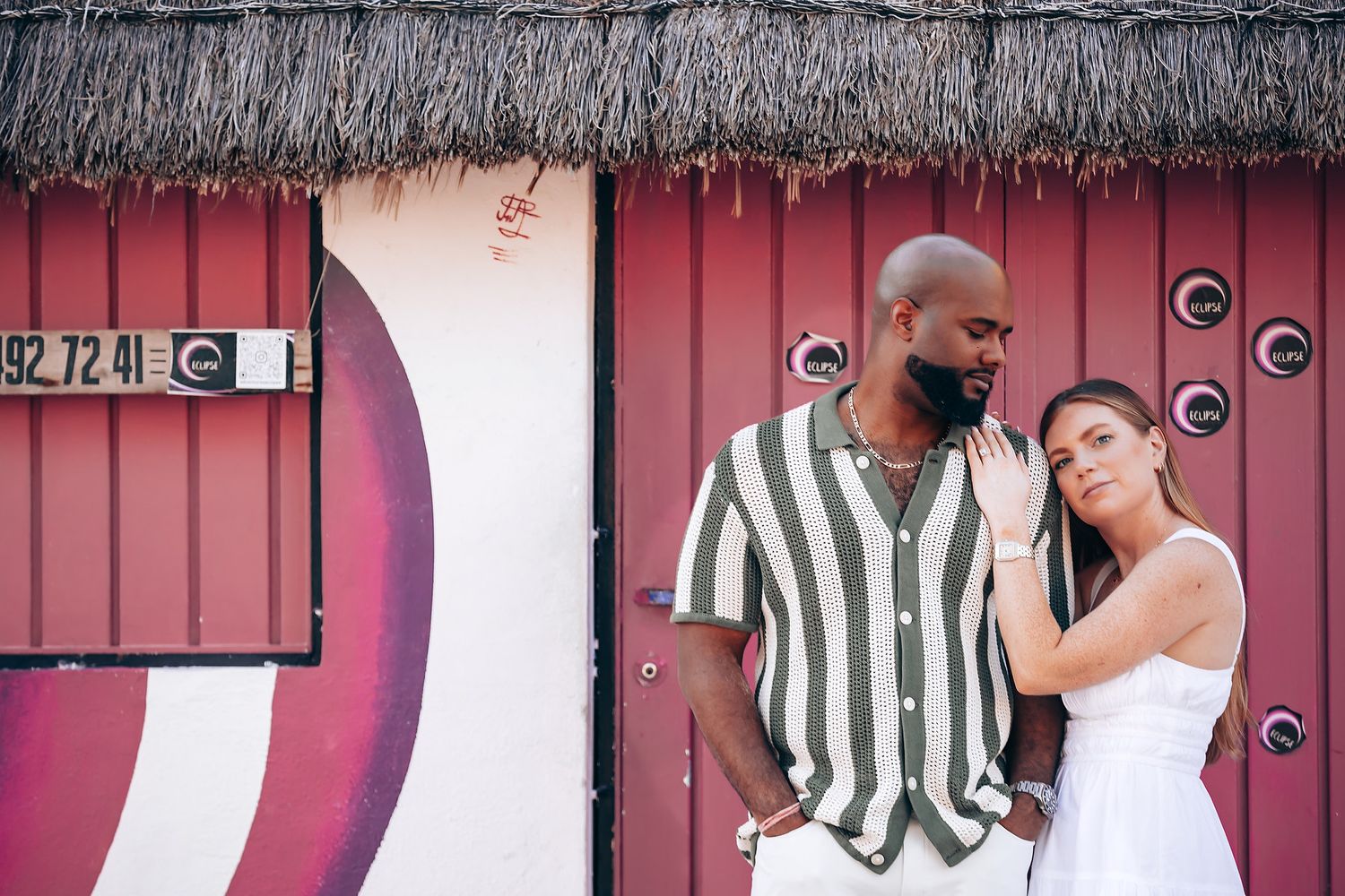A couple poses romantically in front of a red building with thatched roof, man in striped shirt, woman in white dress.