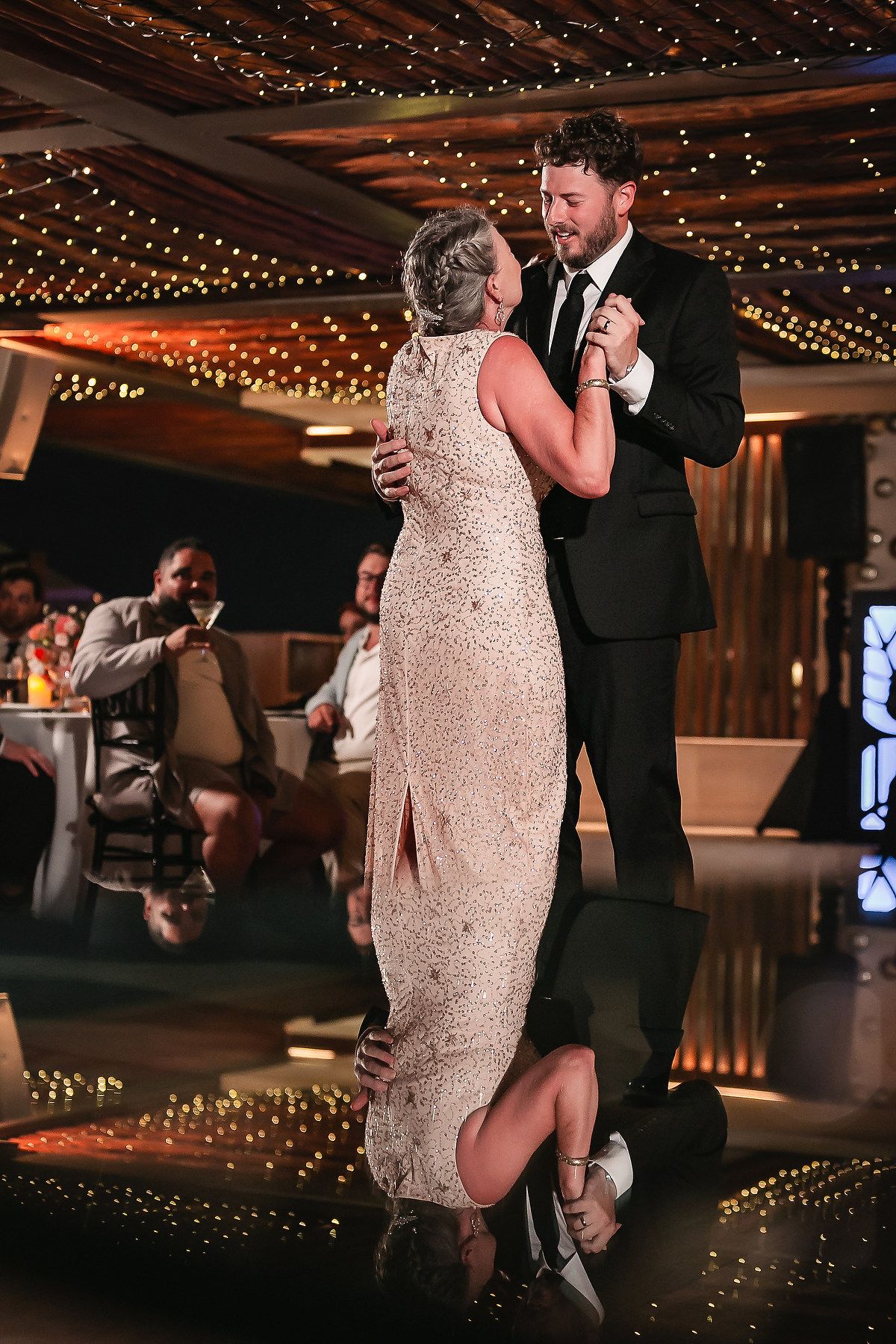 Wedding couple dancing on a reflective floor under warm string lights at their reception.