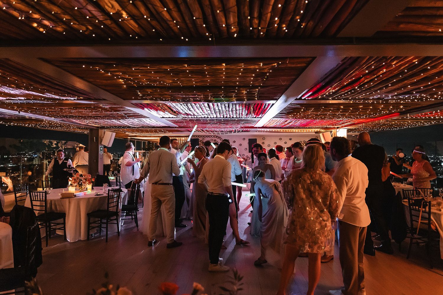Panoramic view of a wedding reception with string lights and dancing guests under a palapa roof.