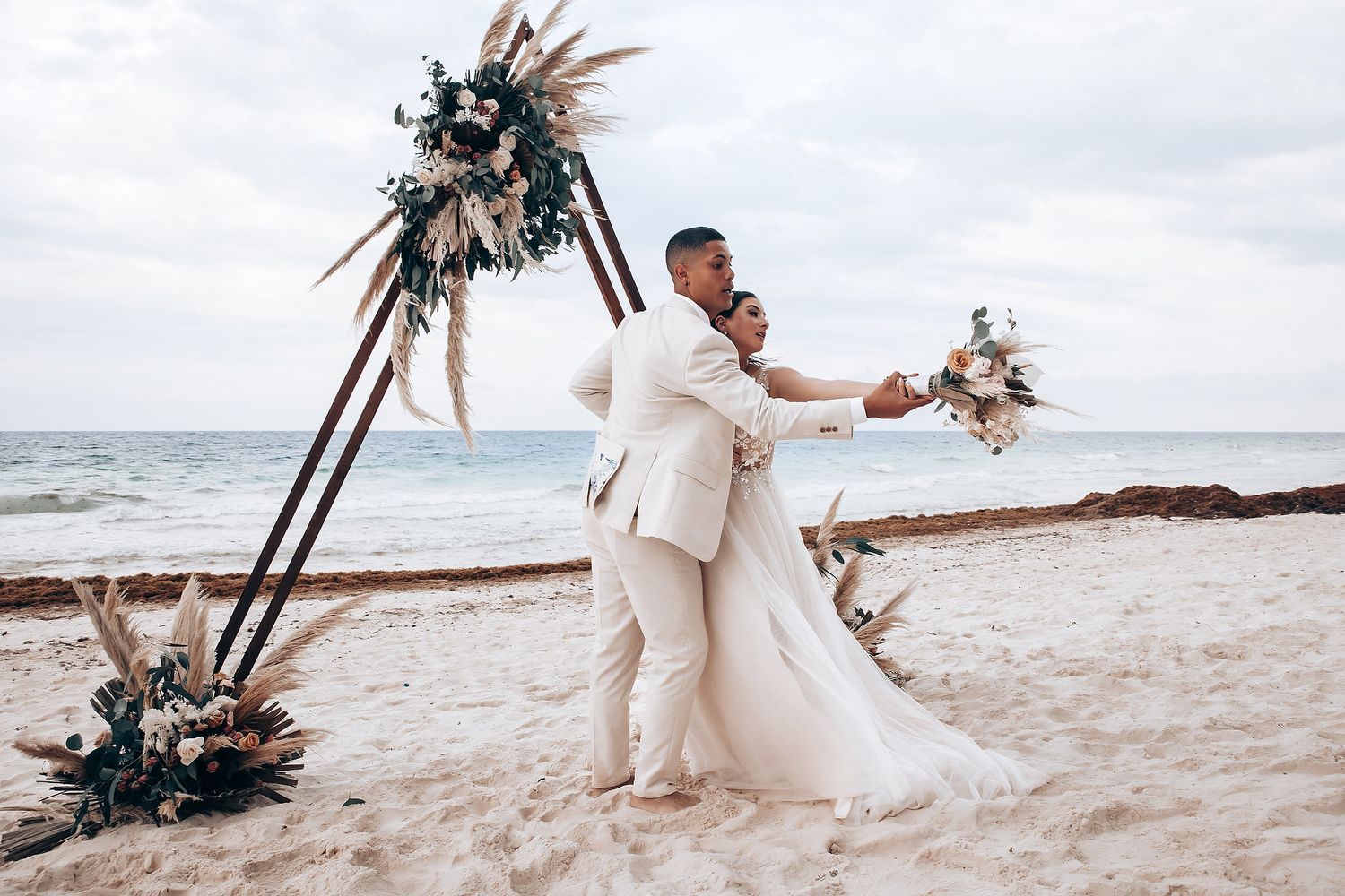 Beach wedding ceremony with wooden arch decorated in dried flowers and pampas grass.