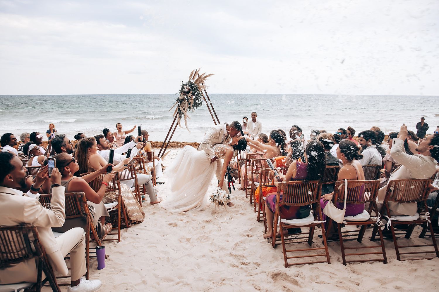 A beach wedding ceremony with guests seated beneath a floral arch by the ocean.