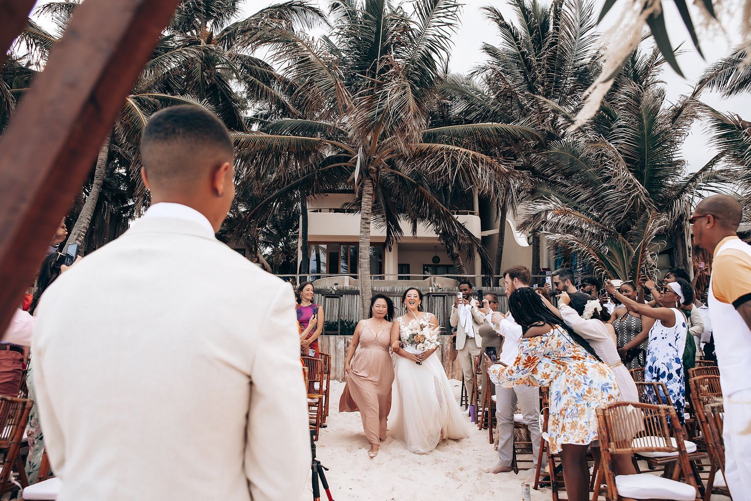 Beachside wedding ceremony setup with white chairs and palm trees along a tropical shoreline.