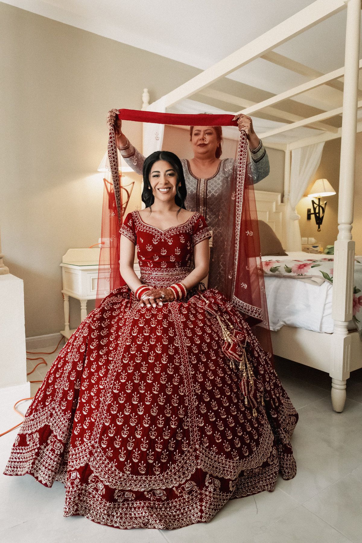Indian bride in red lehenga smiling as woman drapes red dupatta over her in elegant bedroom.