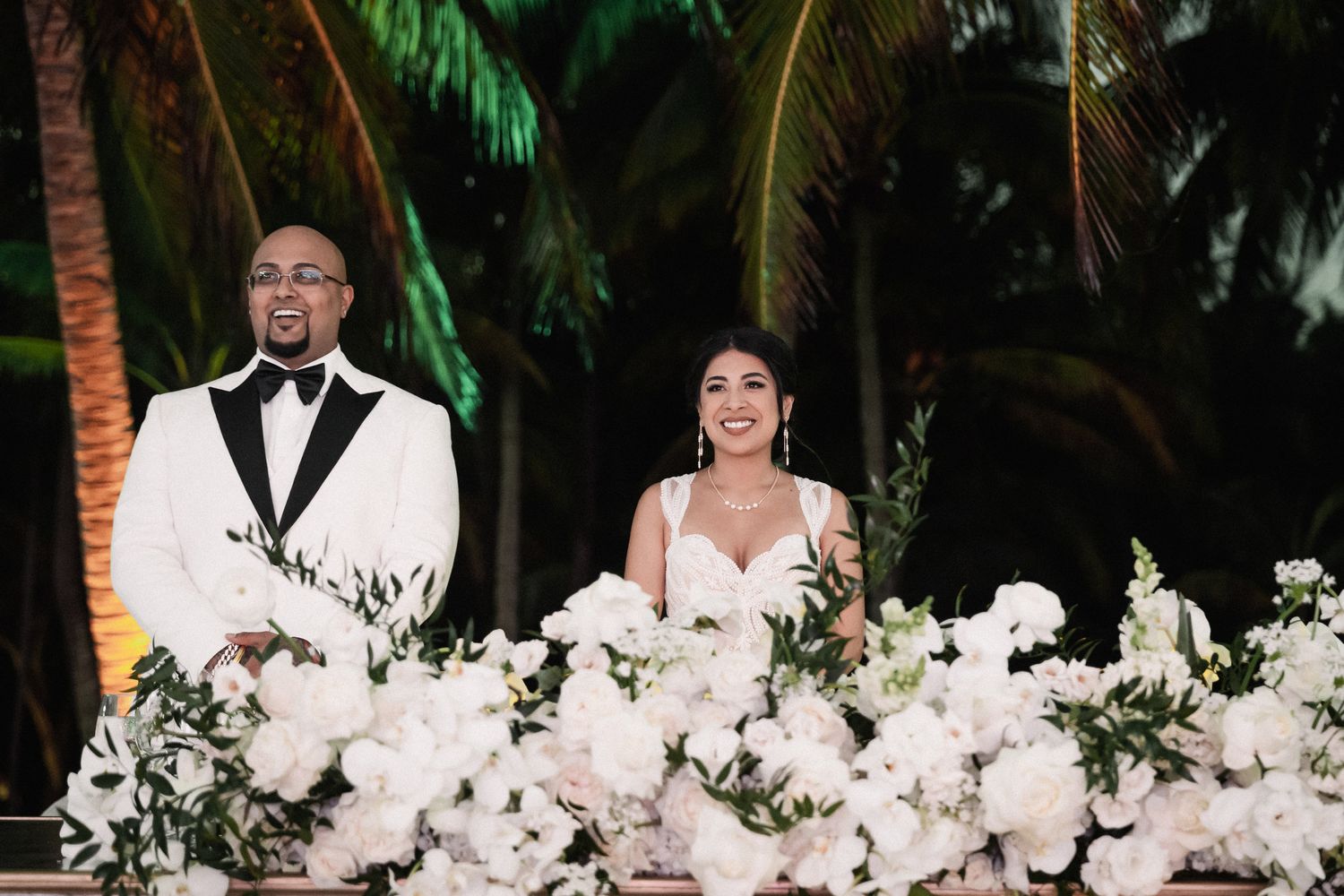 Newlyweds smile at sweetheart table adorned with elegant white floral arrangements at beach wedding.