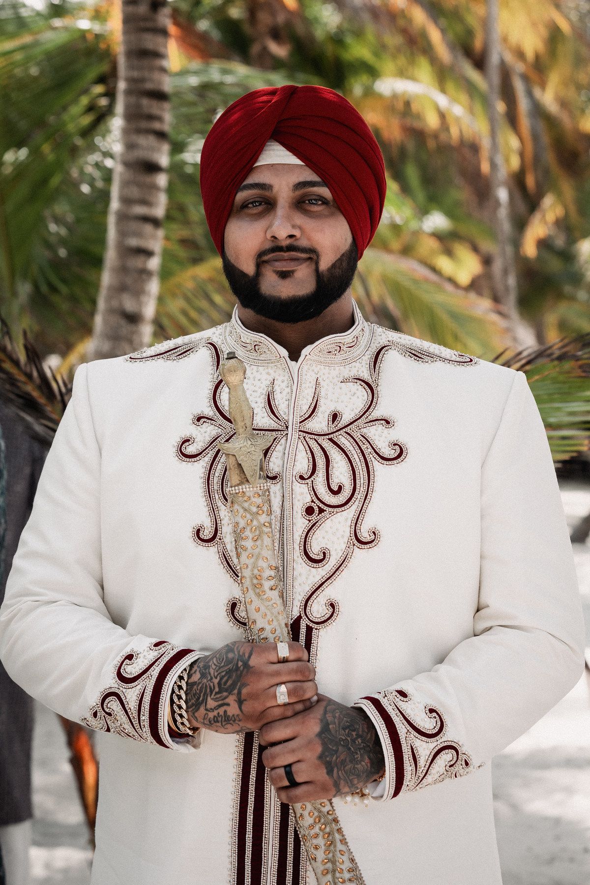 Sikh groom in white embroidered sherwani and red turban holds ceremonial sword at tropical wedding.