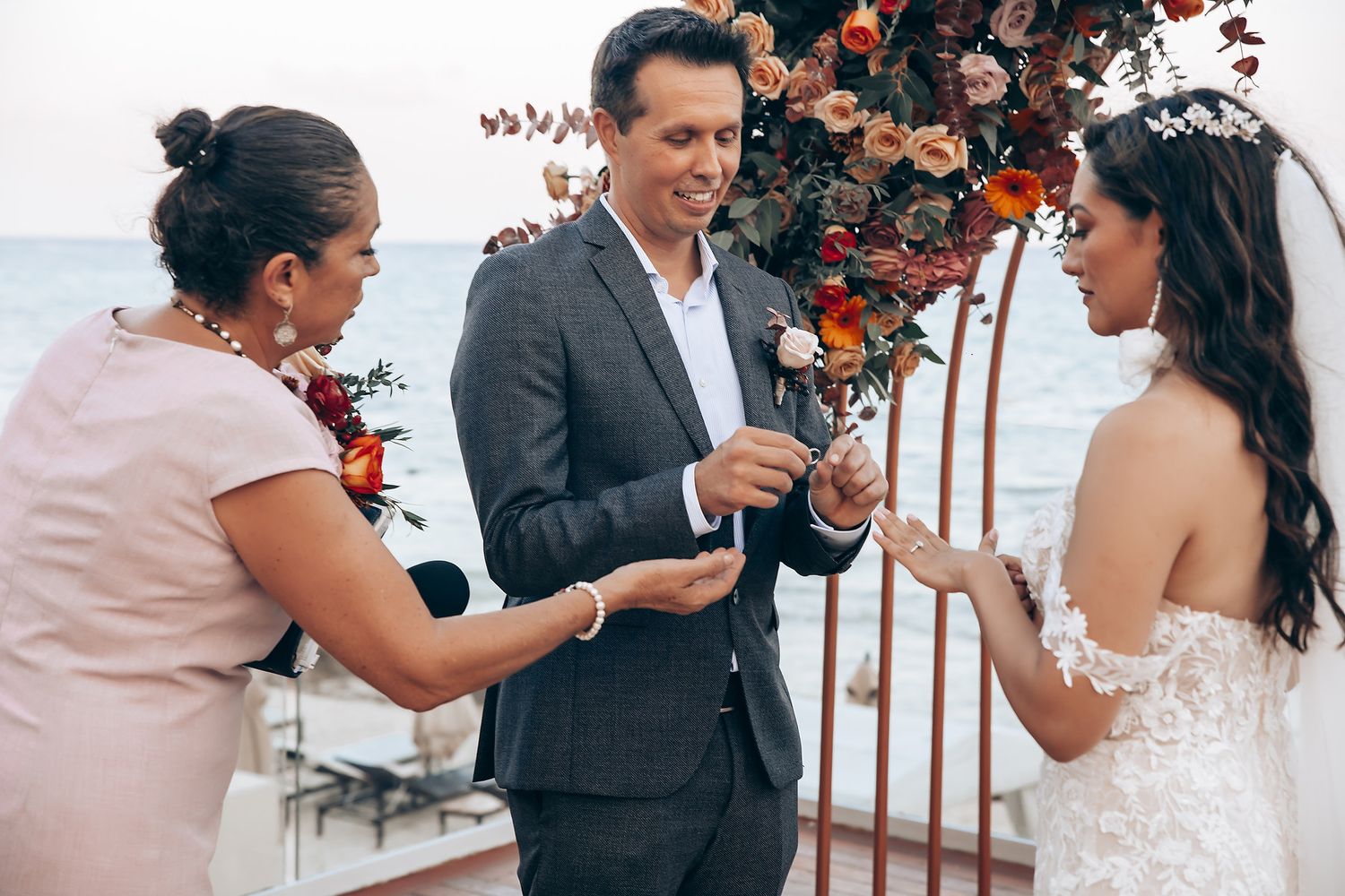 A wedding ceremony moment under a floral arch by the ocean during ring exchange.