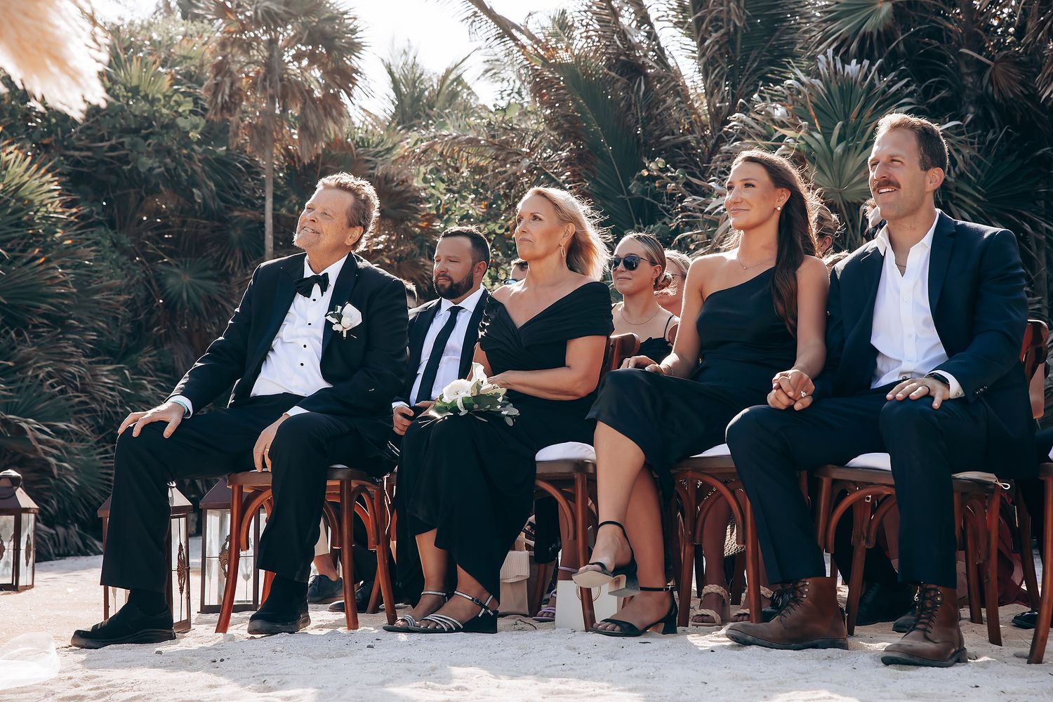 Wedding guests seated in wooden chairs during outdoor ceremony with tropical foliage.
