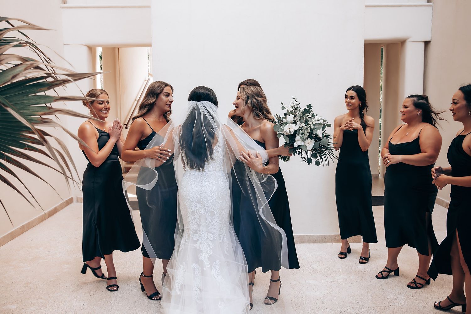 A group of women in black dresses and one in a white wedding gown share joyful moments at a wedding celebration.