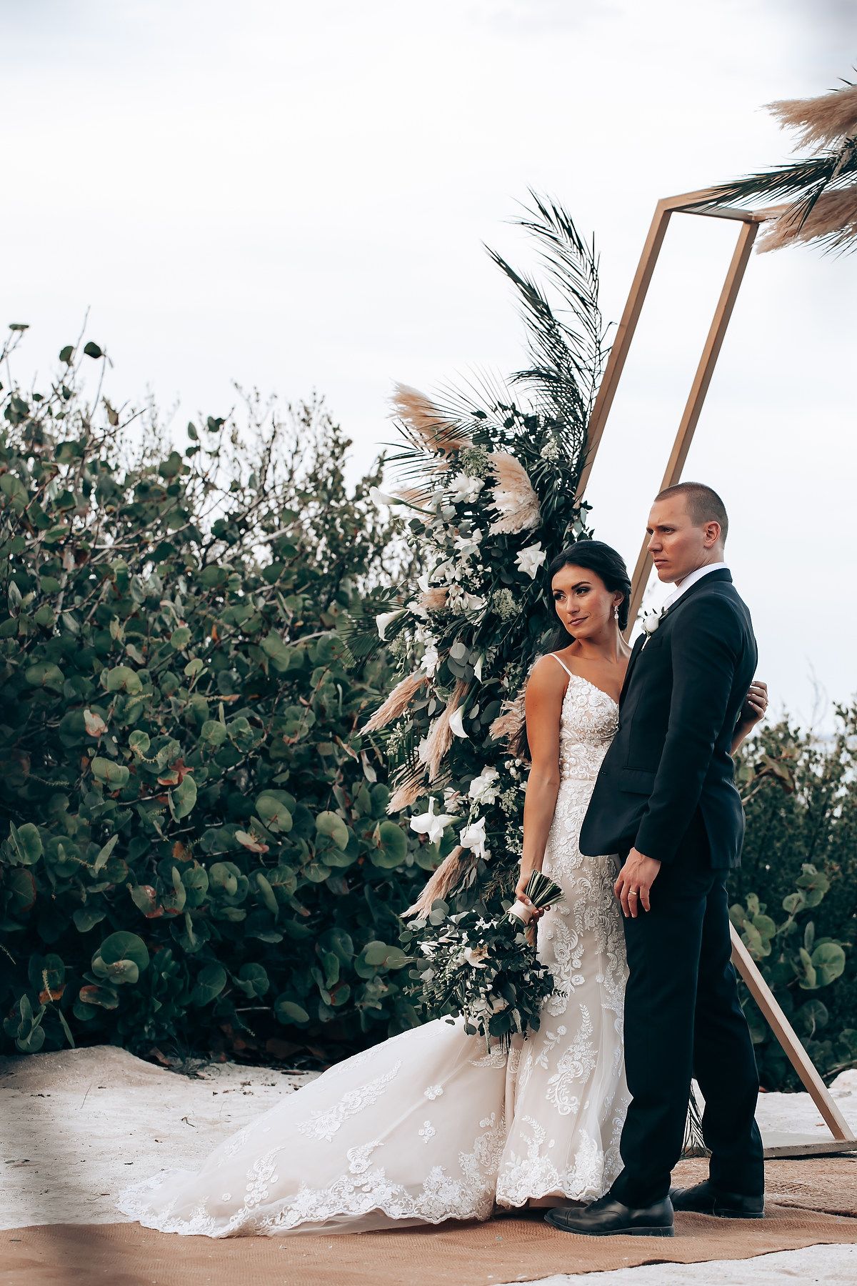 A wedding couple stands together on a beach deck surrounded by lush tropical foliage.