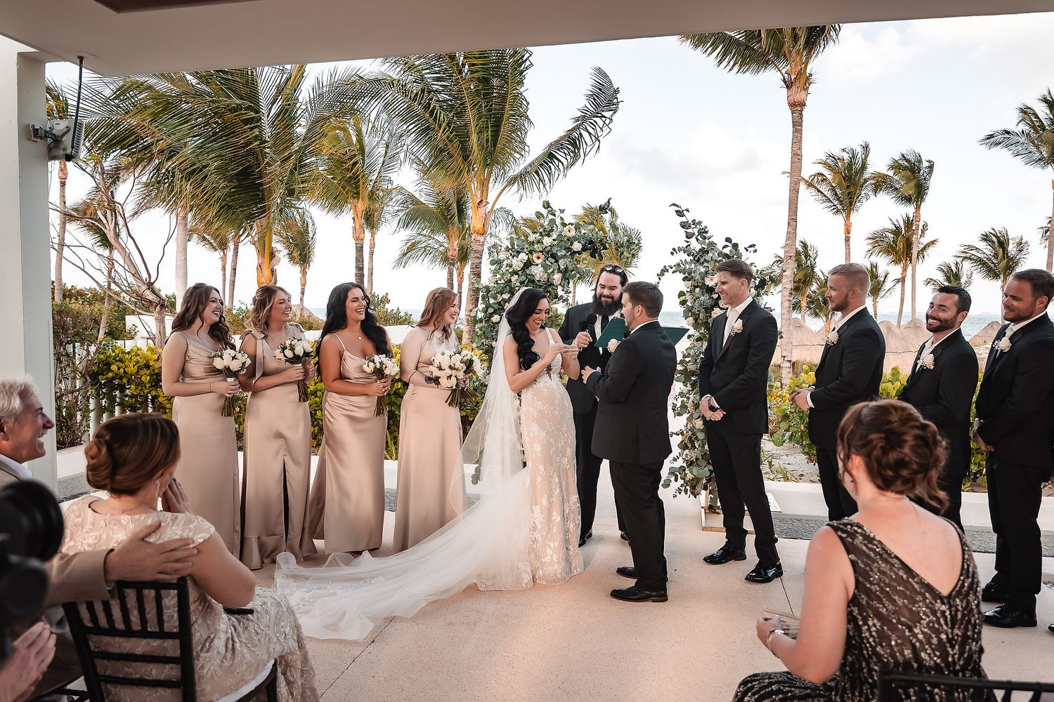 Outdoor wedding ceremony with bridesmaids in champagne gowns and groomsmen in black suits at a tropical beachside venue.