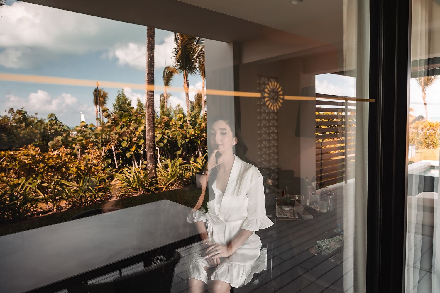 Woman in white robe sitting by glass door with tropical garden reflection.