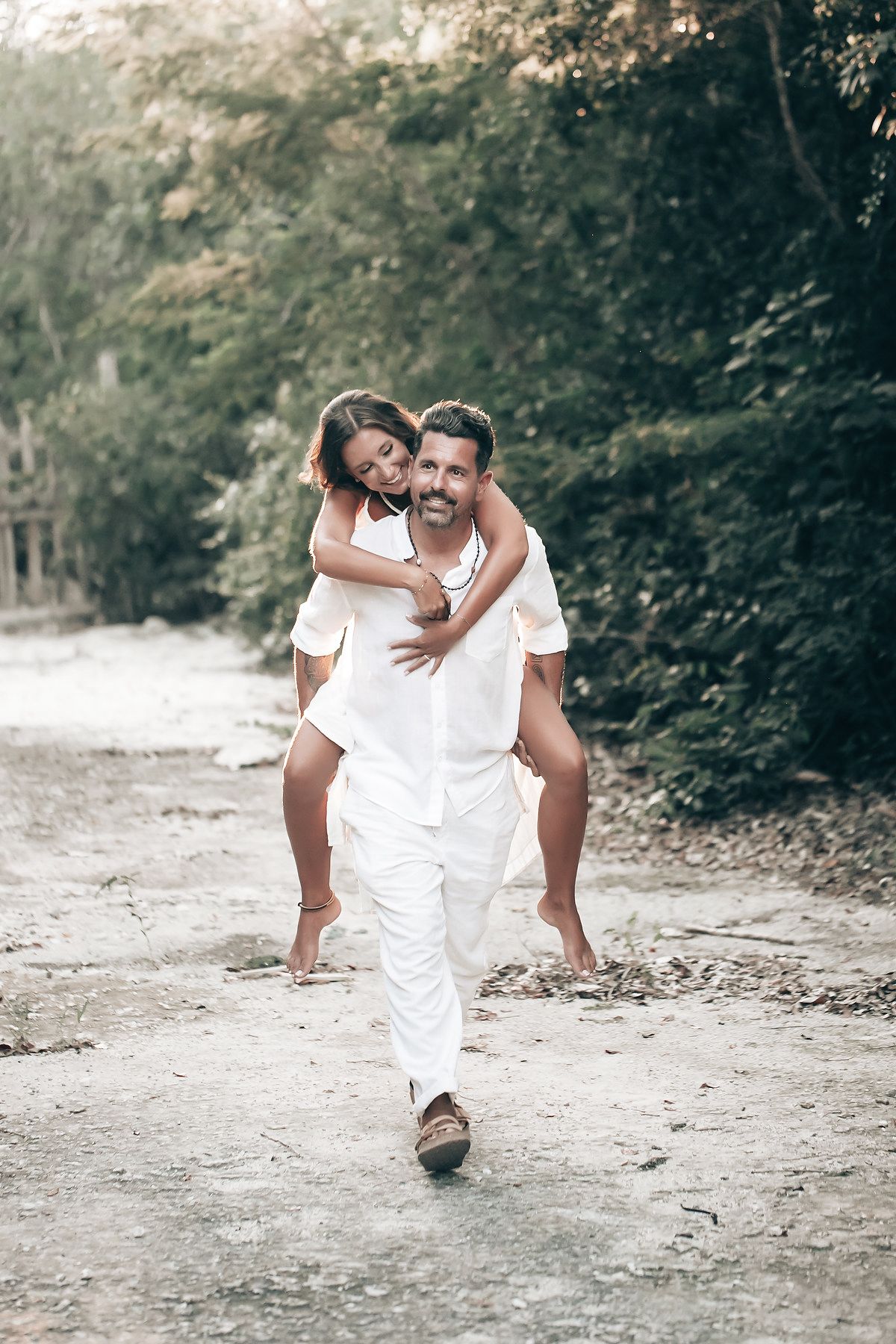 A couple in white clothing embrace and walk together down a dirt path in a forested area.