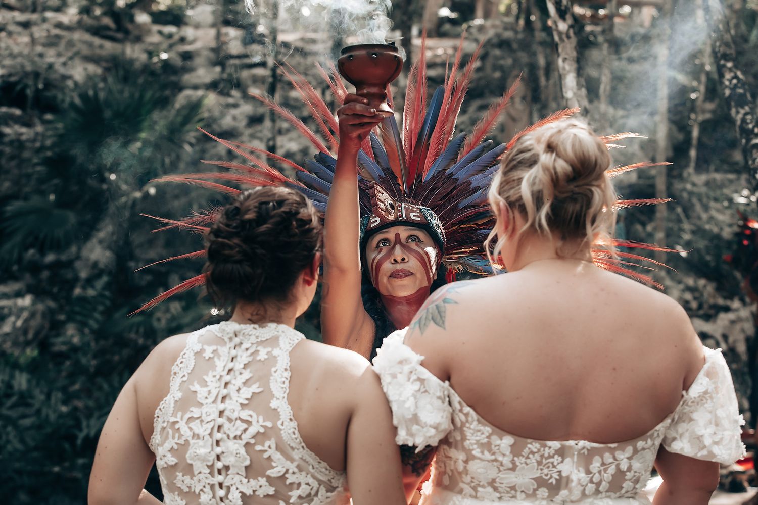 The image shows two women in wedding dresses standing together, with one wearing a colorful feathered headdress.