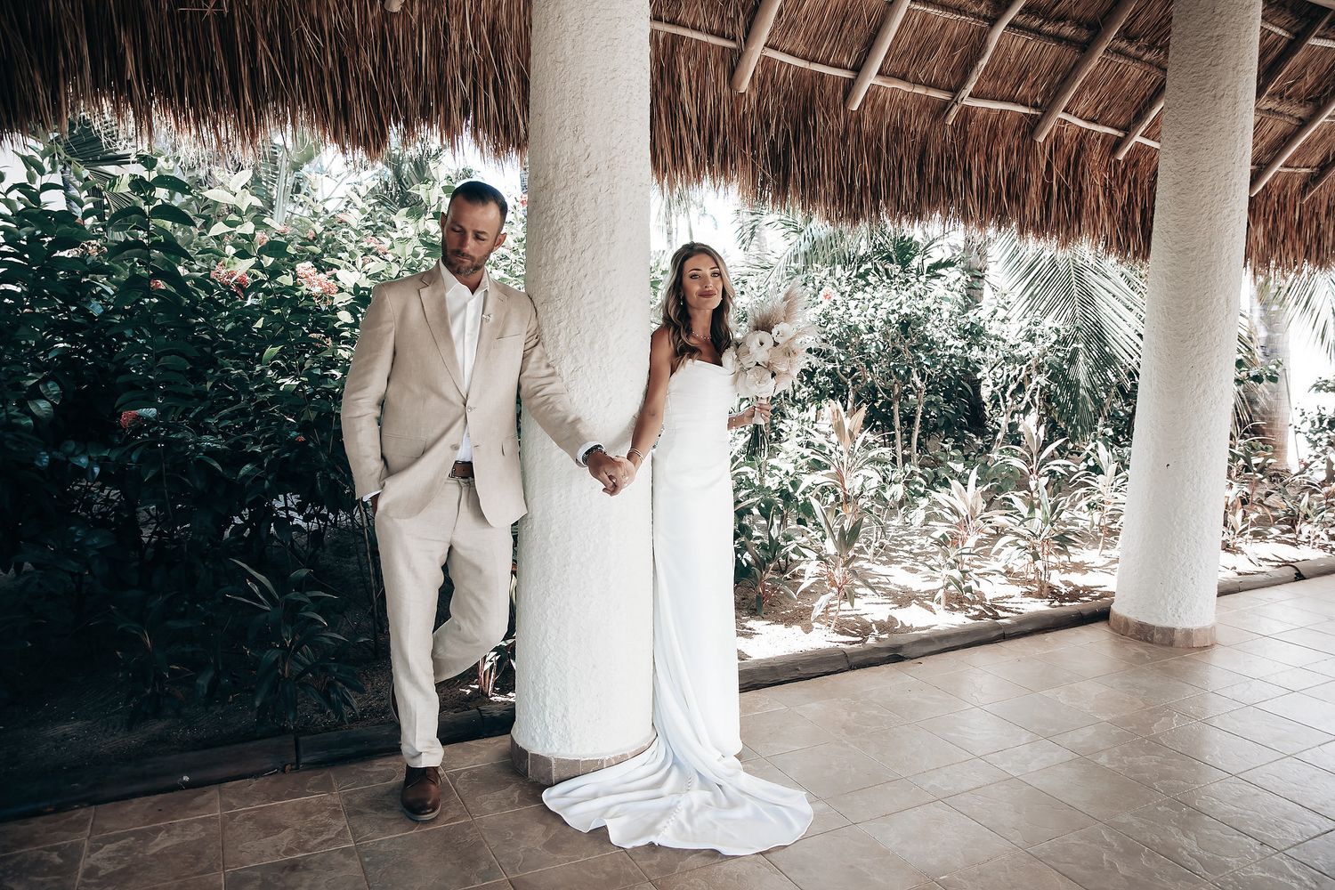 A couple in formal attire stand together on a patio surrounded by greenery.