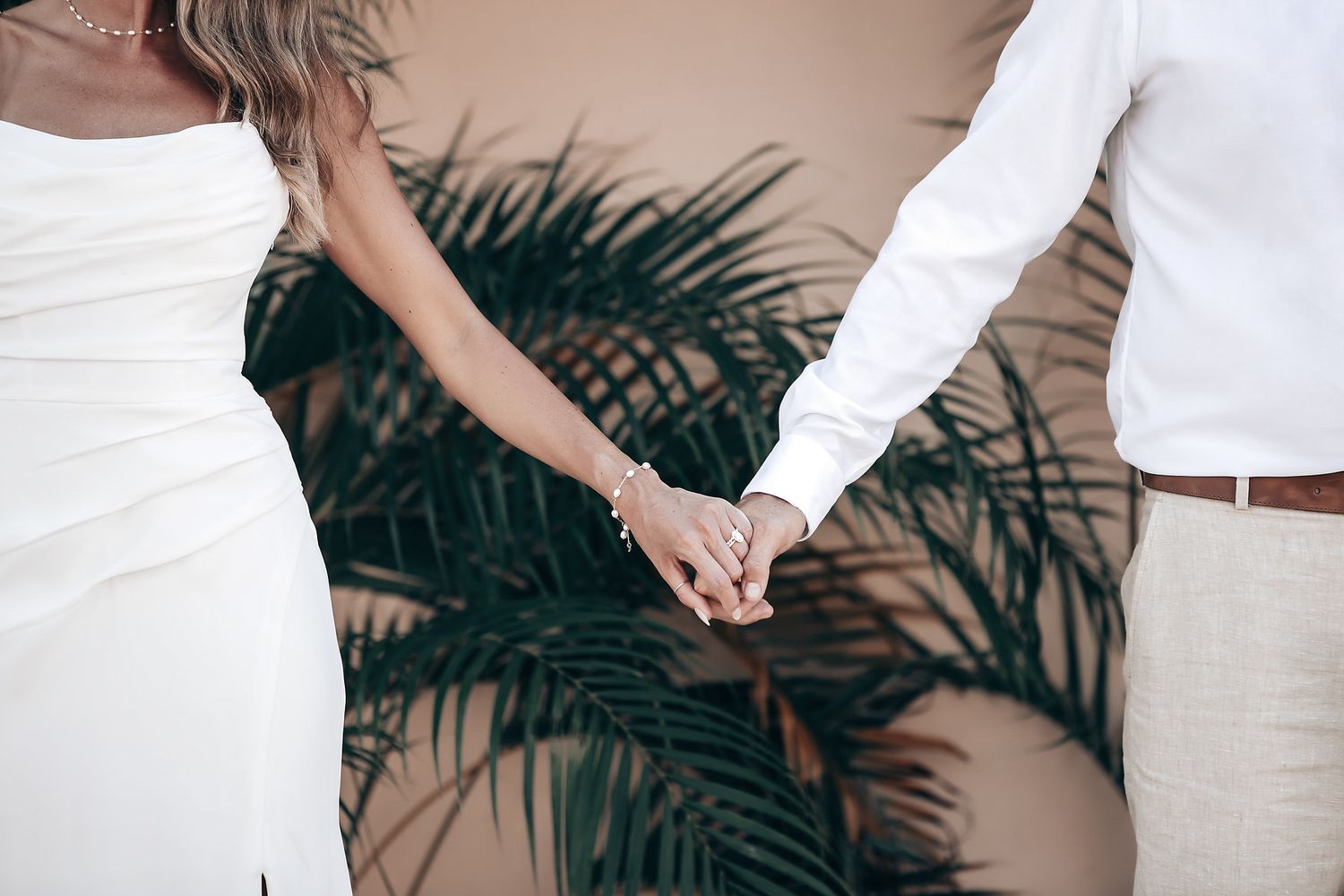 A bride and groom hold hands and embrace in an outdoor wedding setting with palm trees.