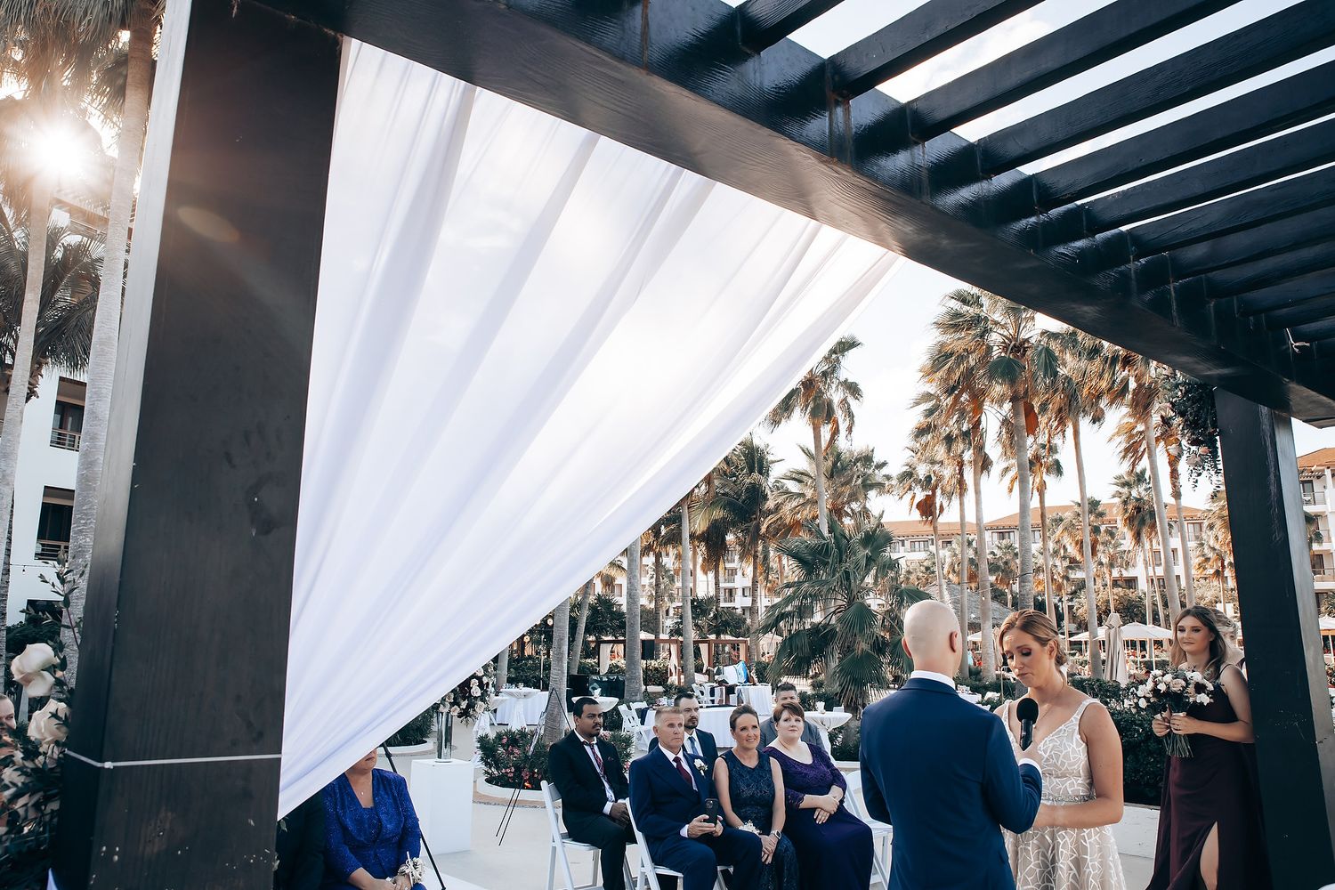 A wedding ceremony taking place outdoors, with guests seated and a white arch or structure visible in the background.