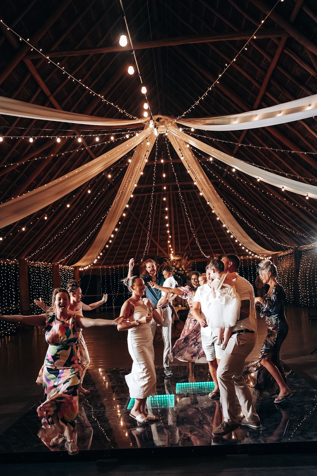 Wedding guests dance under draped fabric and string lights in a rustic barn reception venue at night.