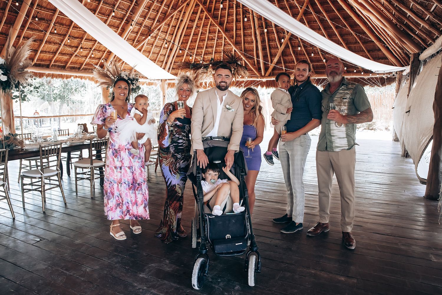 Wedding guests gathered under a large open-air thatched roof pavilion.