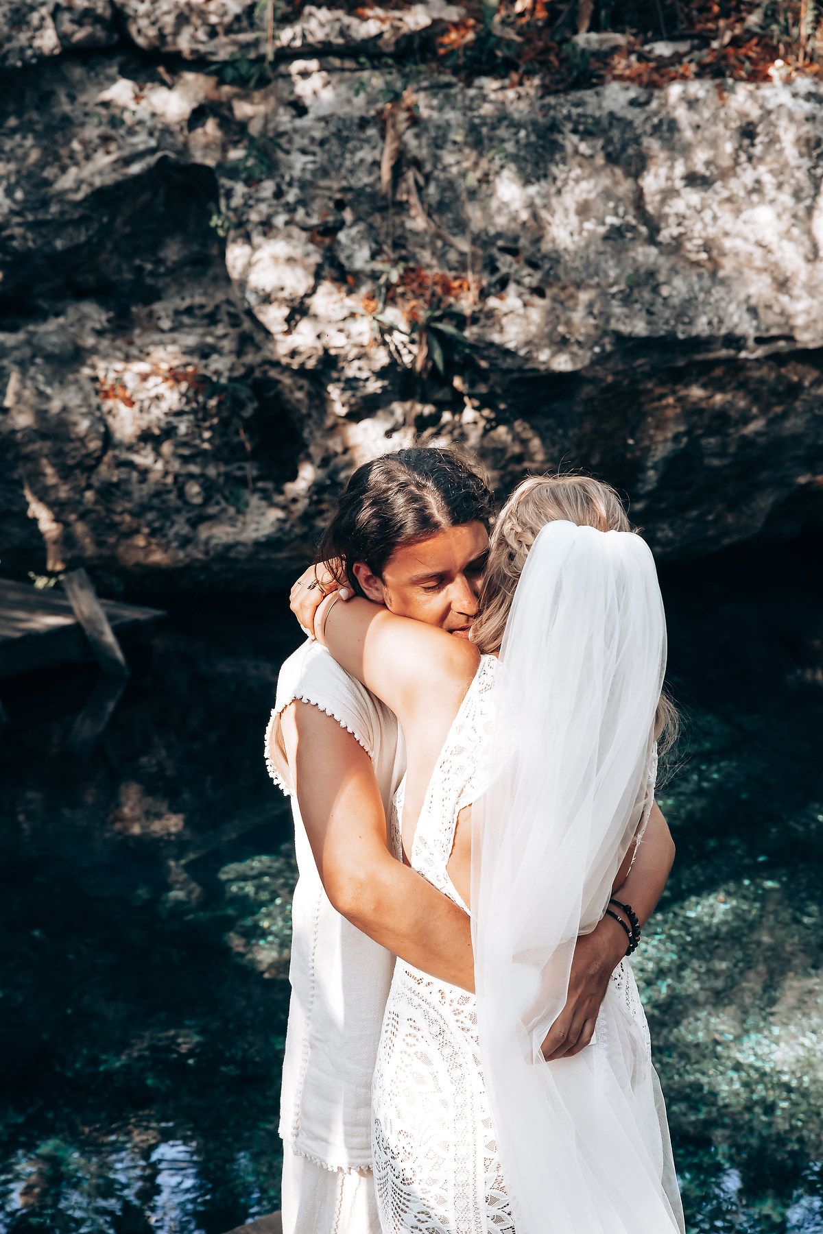 Two people in white wedding attire share an intimate embrace near rocky cliffs and turquoise water.