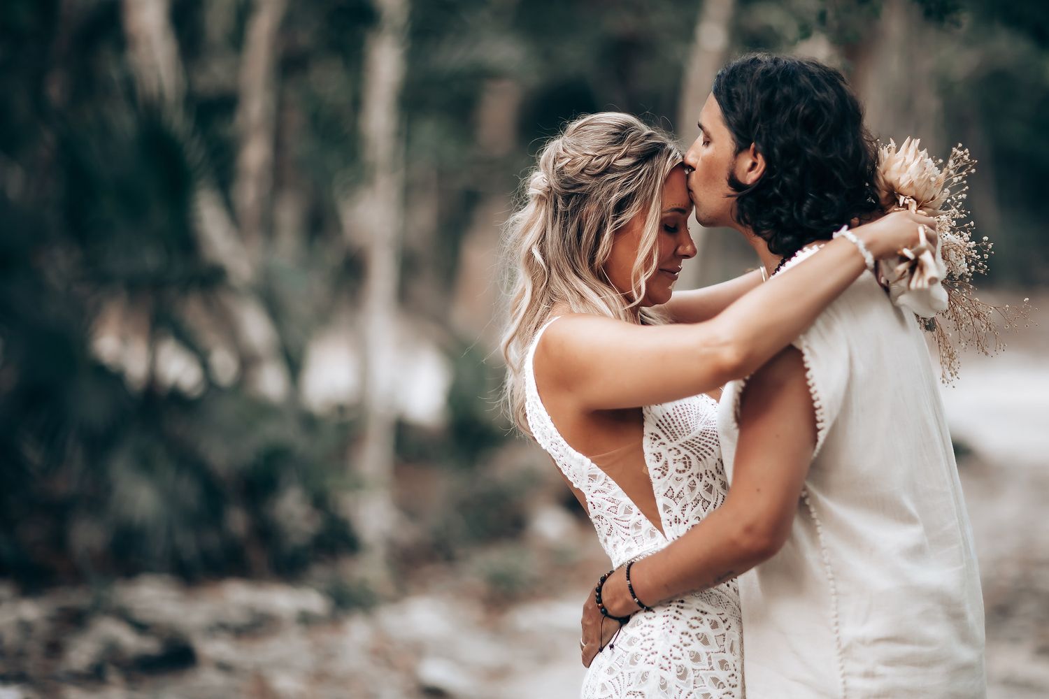 A romantic wedding couple embraces in an intimate moment outdoors with a dreamy forest backdrop.