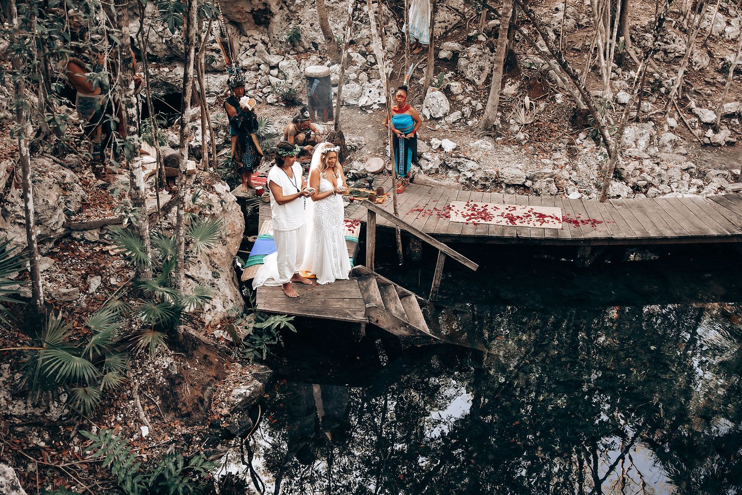 People in white outfits gather on a wooden deck overlooking a reflective pond surrounded by autumn trees.