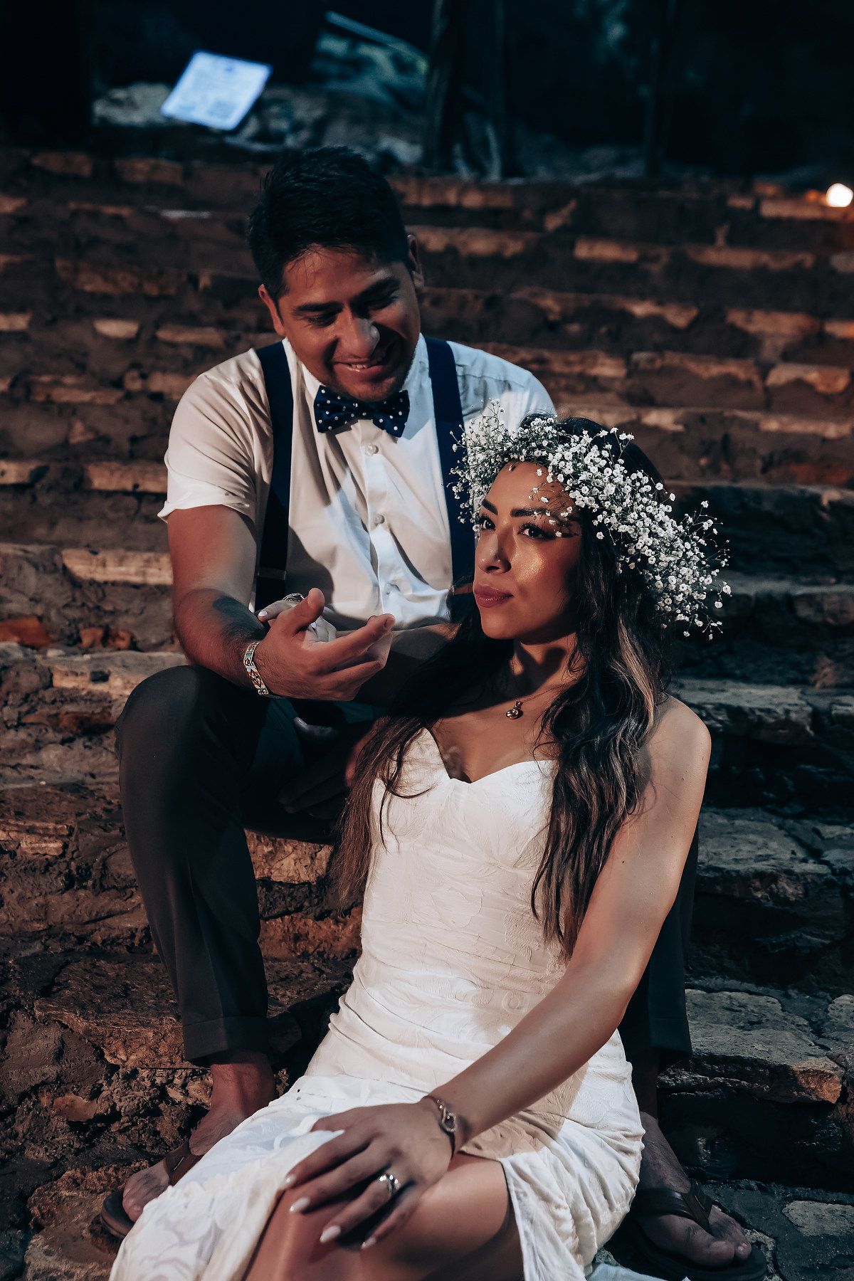 A couple sits together on rustic brick steps decorated with delicate white flowers and romantic lighting.