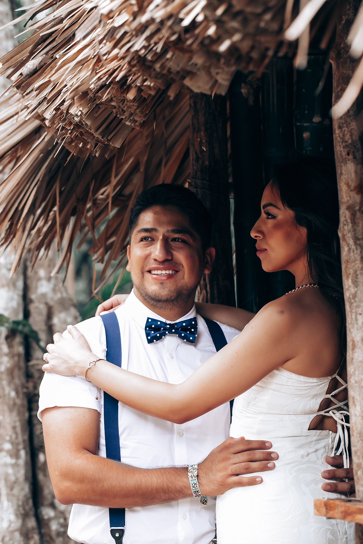A couple shares romantic moments under a thatched palm roof during a tropical beach wedding celebration.