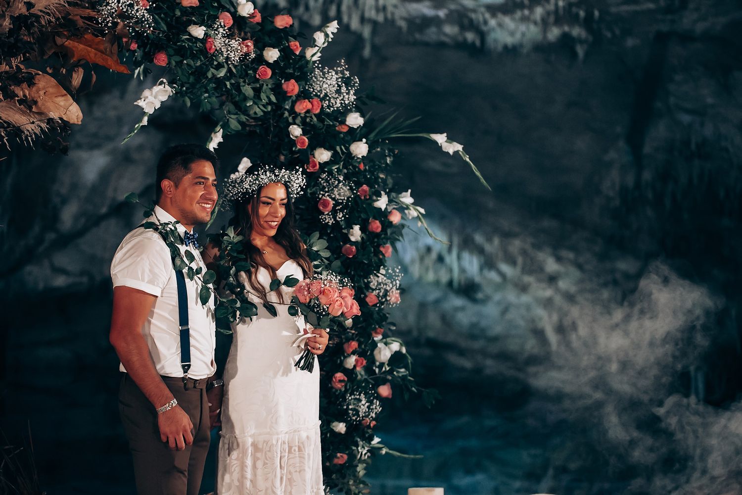 Romantic wedding couple poses under floral arch with coral and white flowers against dramatic dark background.