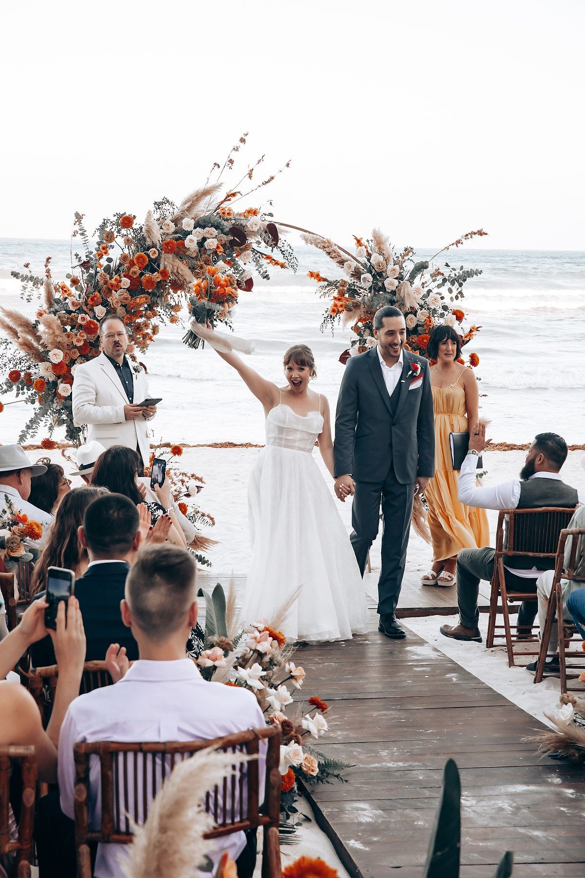 Beachside wedding celebration with coral floral arrangements and wooden chairs.