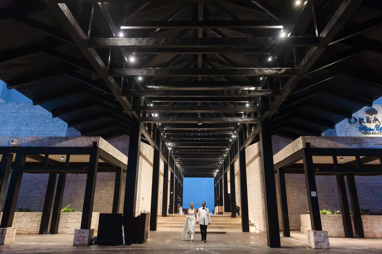 Elegant outdoor venue corridor with black steel beams, recessed lighting, and couple walking at night.