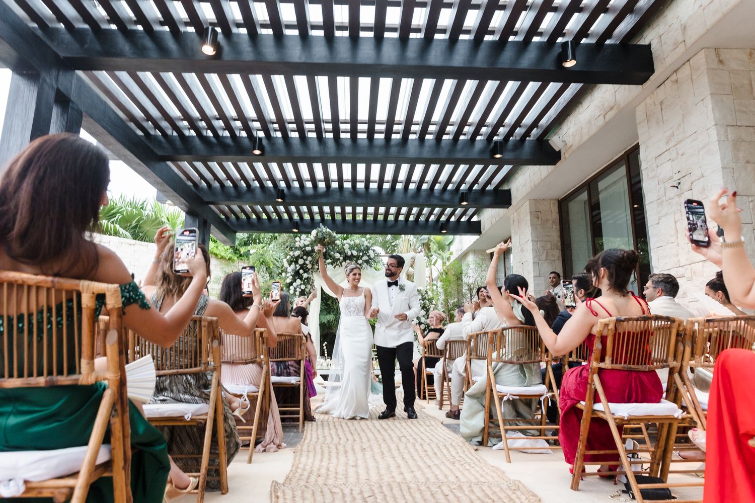 Newlyweds celebrate their outdoor wedding ceremony under a pergola, surrounded by cheering guests in elegant attire.