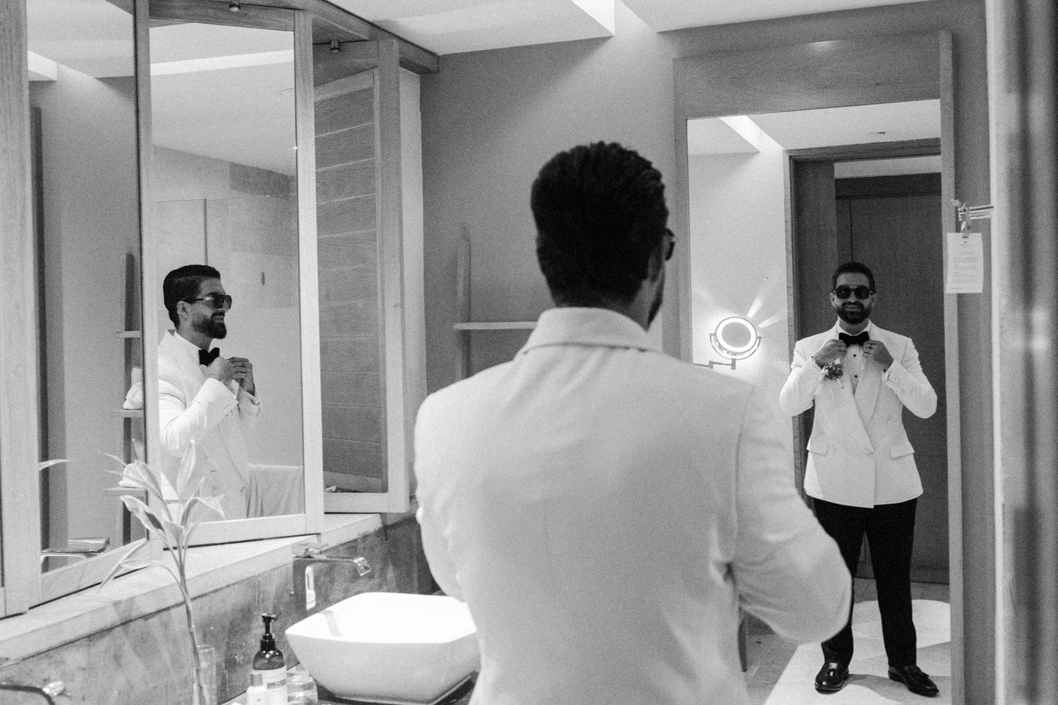 Groom adjusting tie while getting ready in hotel bathroom mirror before wedding day.