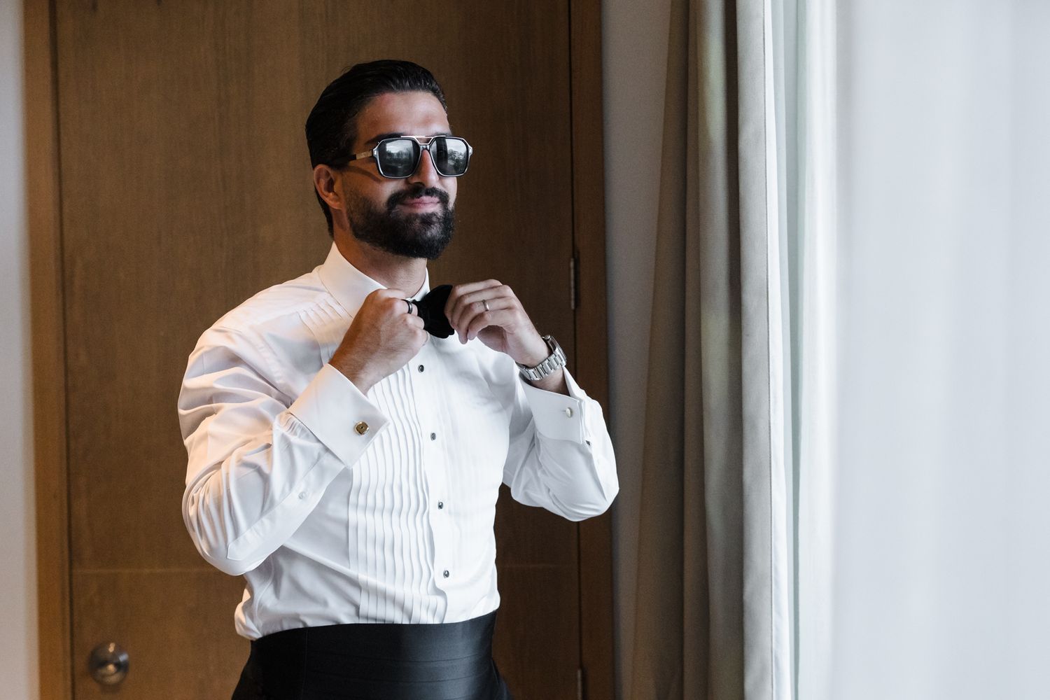 A stylish groom in a white tuxedo shirt and sunglasses adjusts his black bow tie near a window.