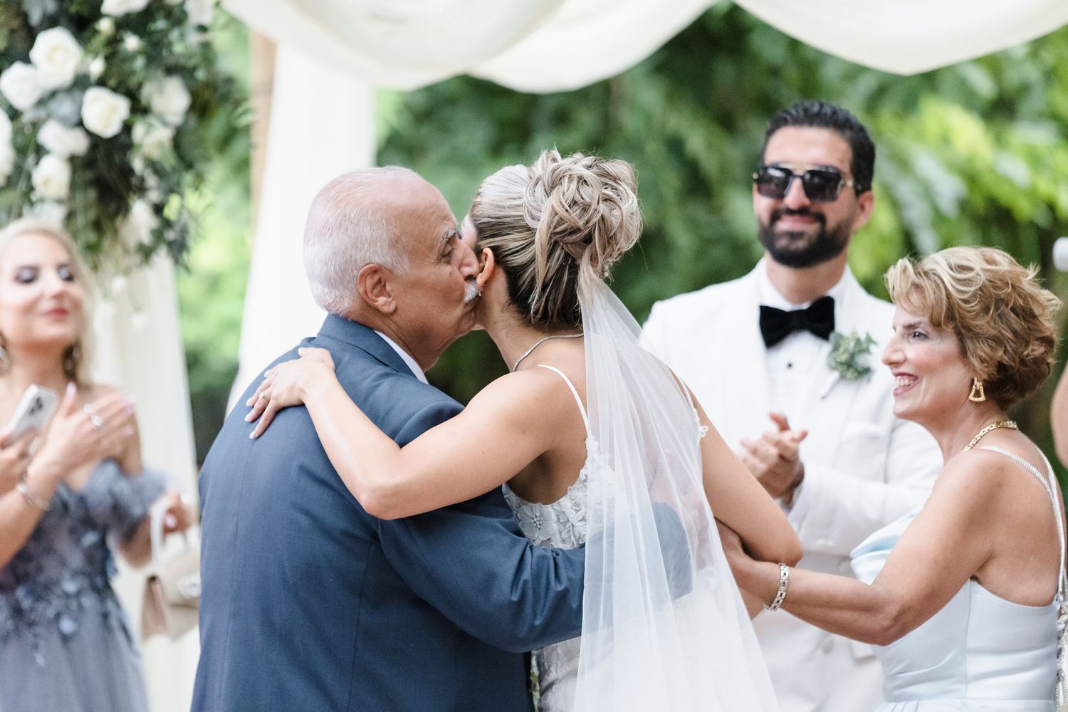 Bride shares a kiss with father at outdoor wedding ceremony while groom and guests celebrate in background.