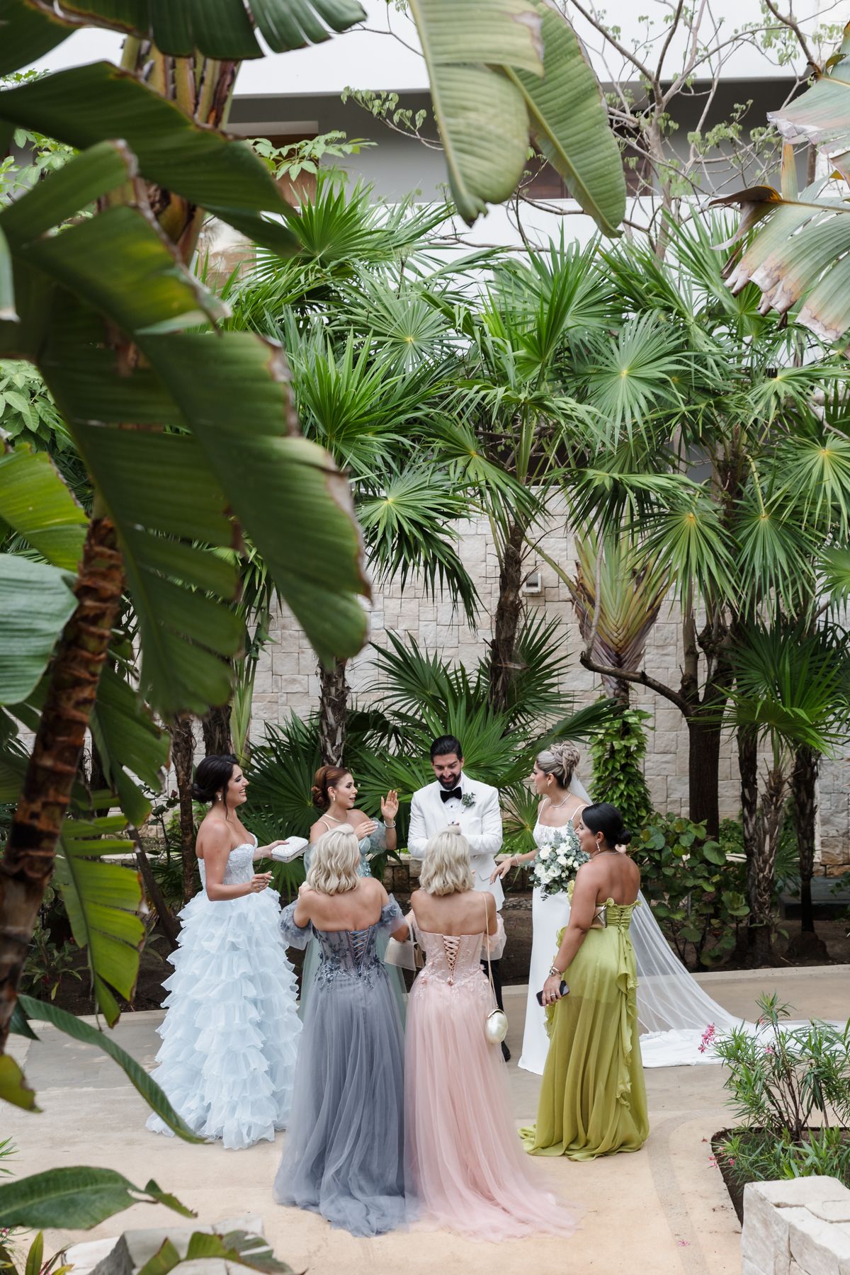 Wedding party gathered with officiant among lush tropical palms at outdoor ceremony.