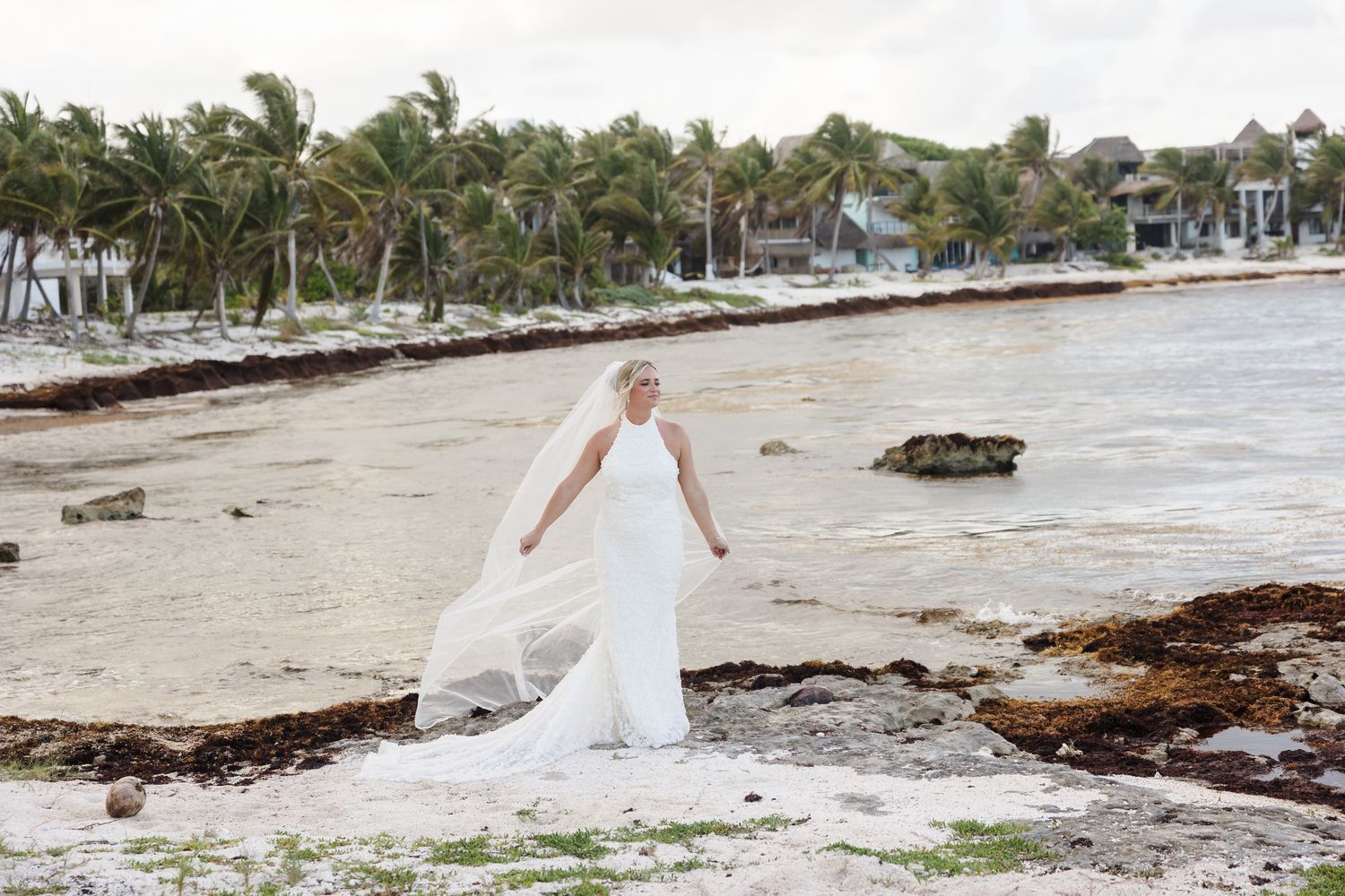 Bride in white gown and veil poses on tropical beach shoreline with palm trees in background.