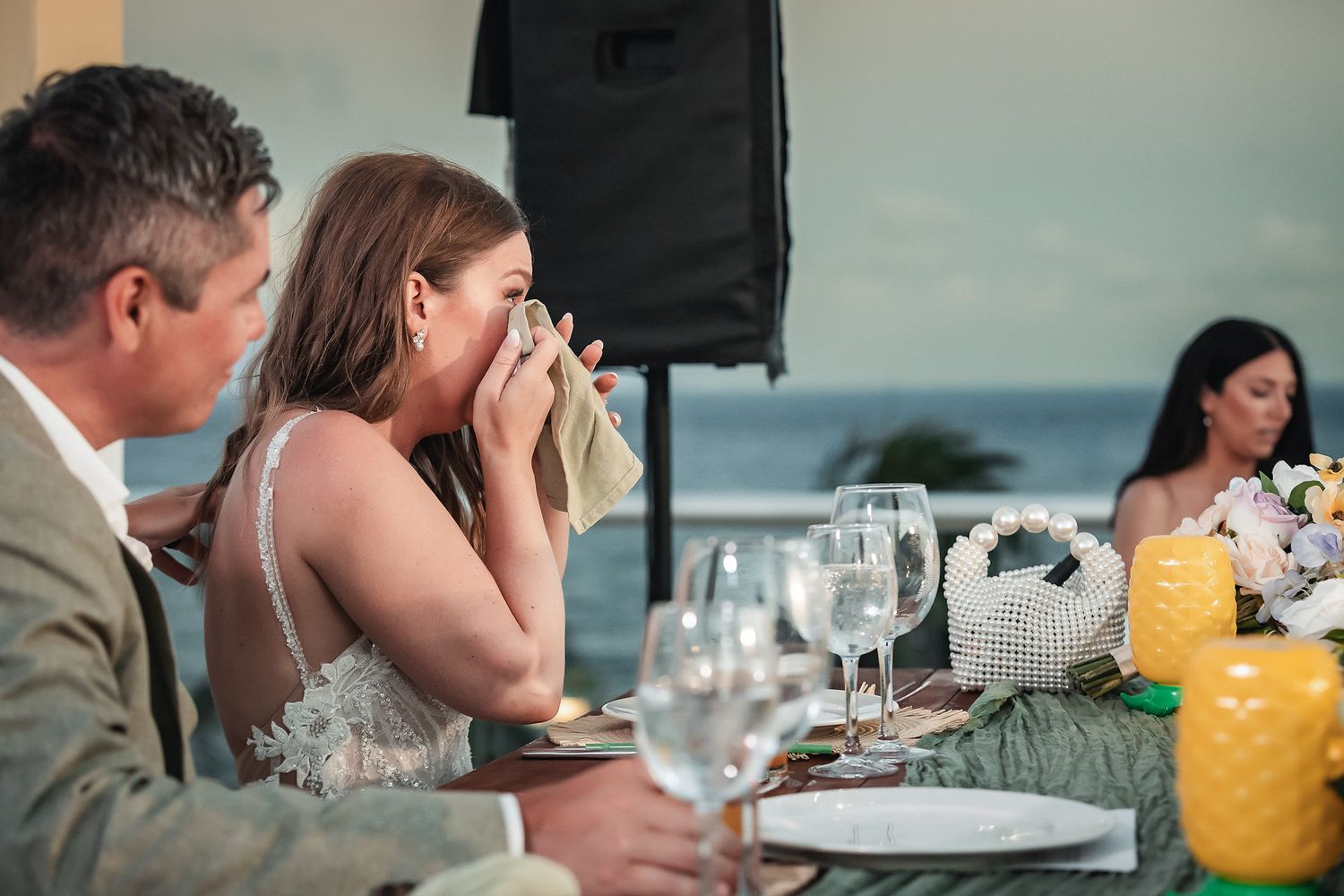 Emotional bride wiping tears at outdoor oceanfront wedding reception dinner table with guests and floral arrangements.