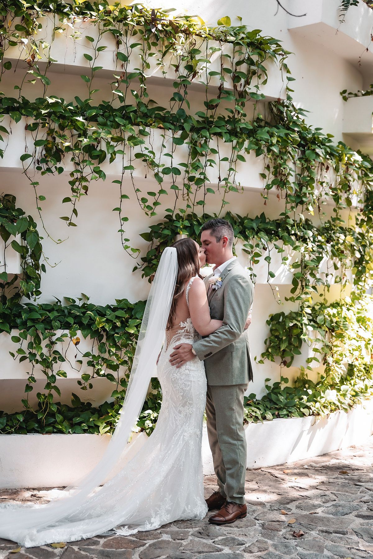 Bride and groom share romantic kiss in front of ivy-covered white wall at outdoor wedding venue.