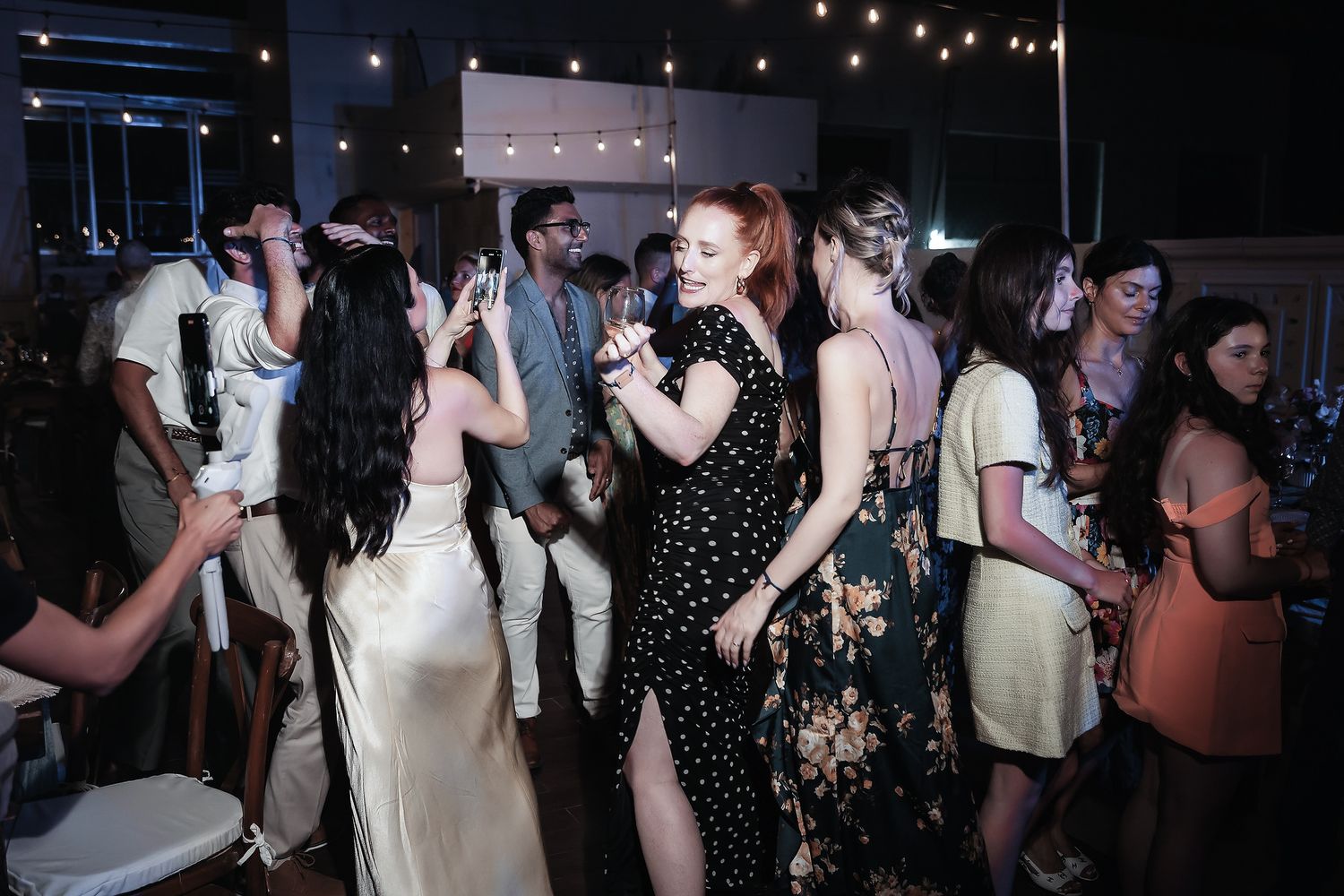 Guests dancing joyfully at an indoor wedding reception with string lights and colorful attire.