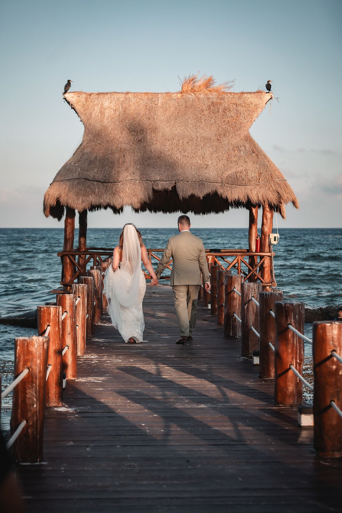 Bride and groom walking hand in hand on a wooden pier toward a thatched palapa overlooking the ocean.