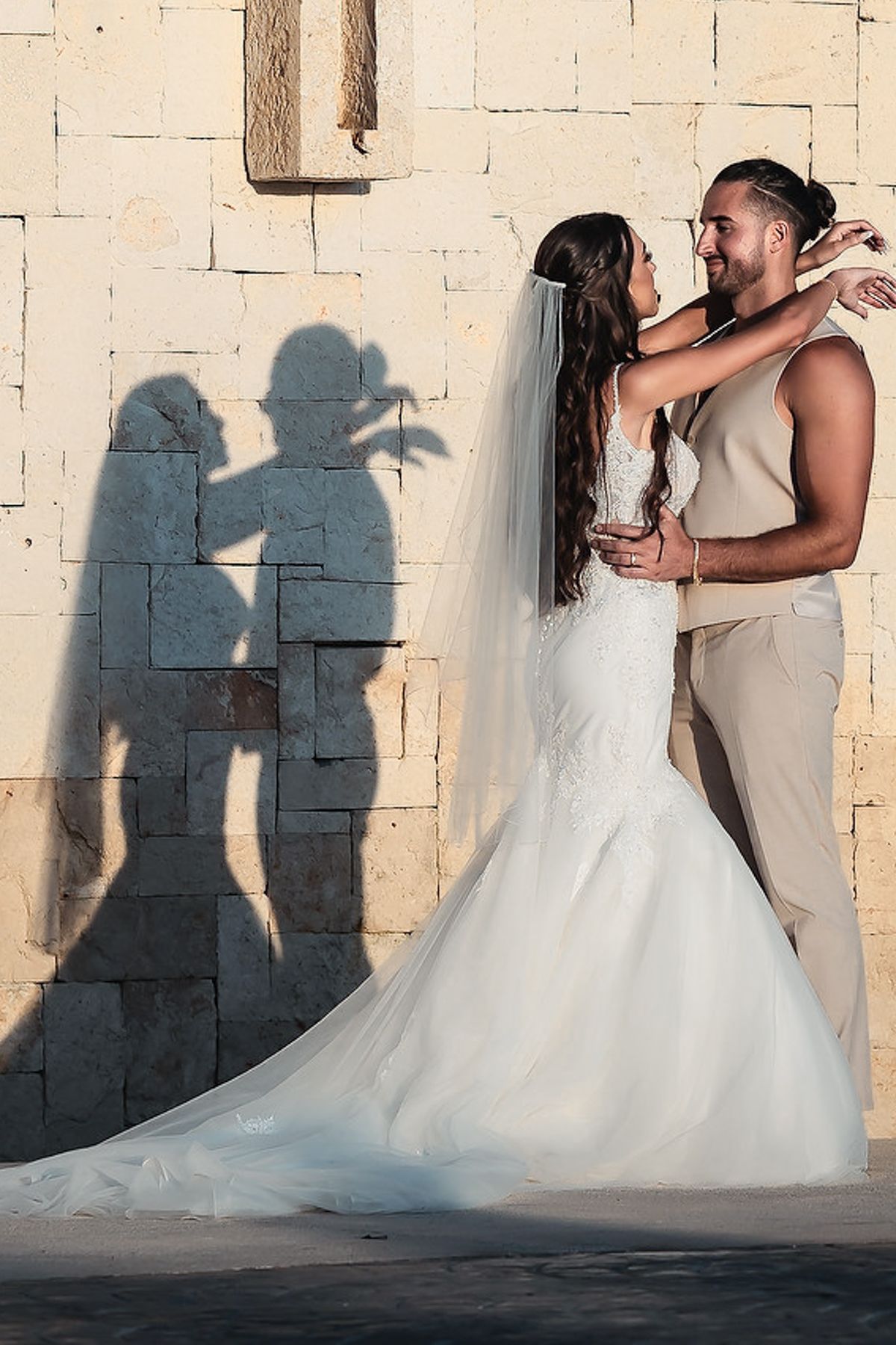 Bride and groom embracing against stone wall with romantic shadows, bride in mermaid dress with veil.