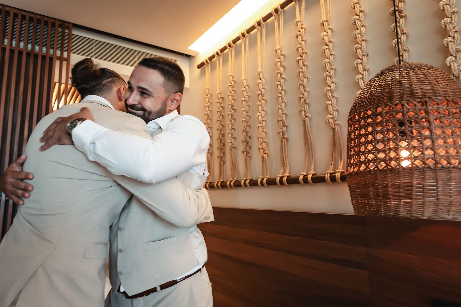 Two men in white shirts share a warm embrace in an elegantly decorated venue with woven wall decor.